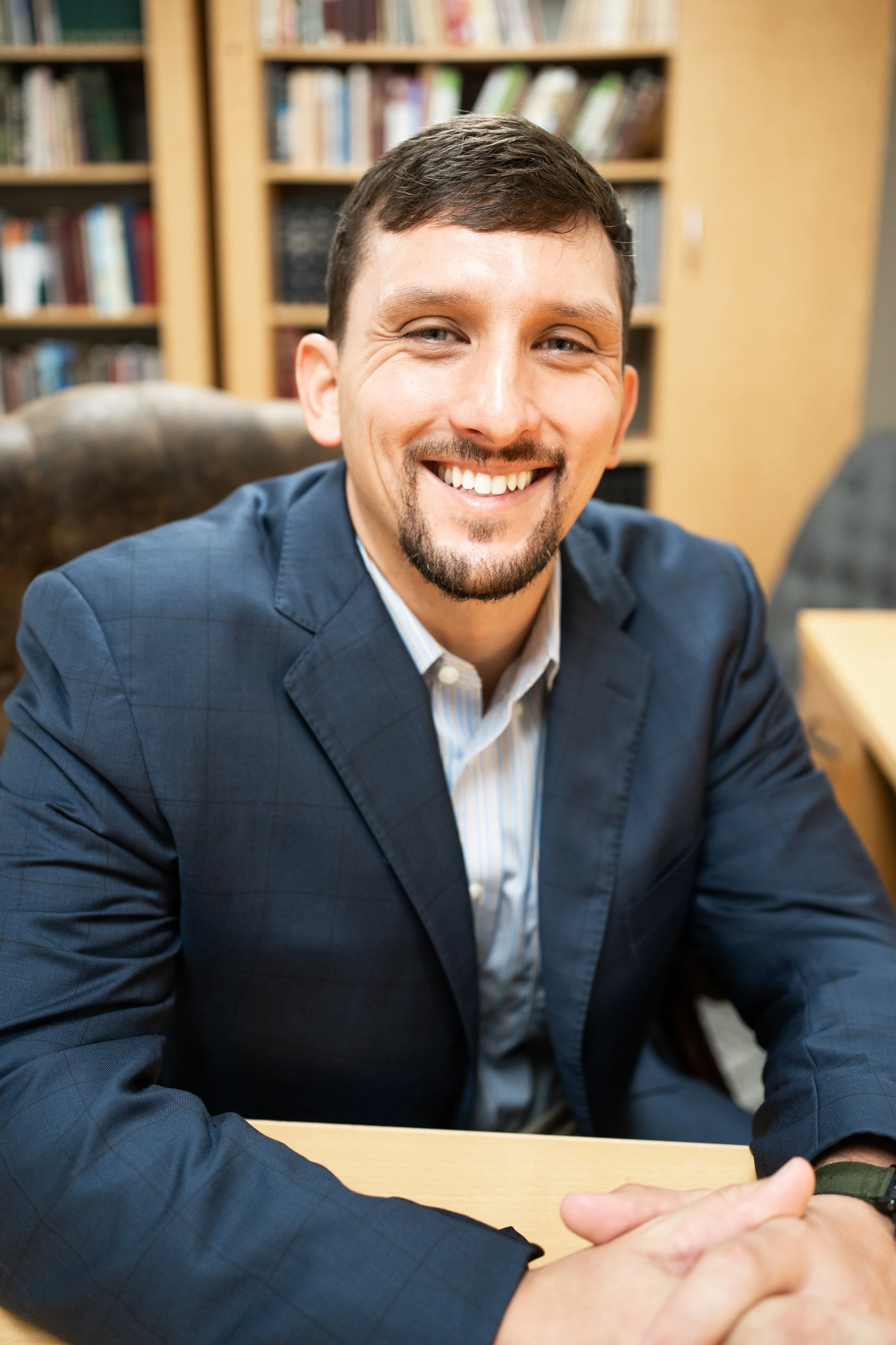 A smiling man with short dark hair and a goatee wearing a blue suit jacket and a white dress shirt, sitting at a desk in front of a bookshelf filled with books.