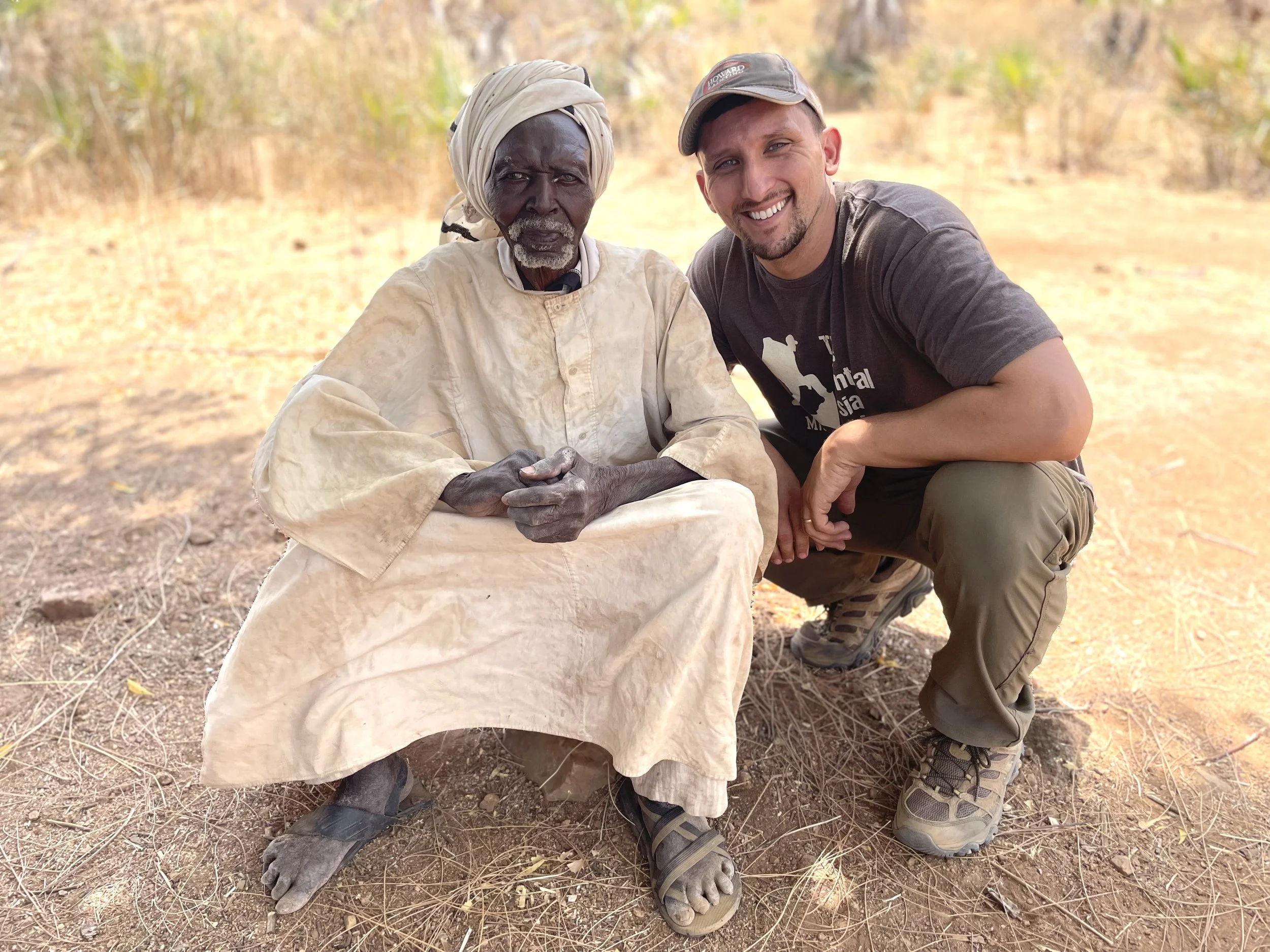 Two men squatting outdoors in a dry, grassy area, smiling for the camera. One man appears to be older, with dark skin, gray hair, wearing a beige traditional outfit and head covering. The other man appears to be younger, with light skin, light hair, wearing a baseball cap and casual clothing. They are surrounded by a yellowish landscape with scattered trees.