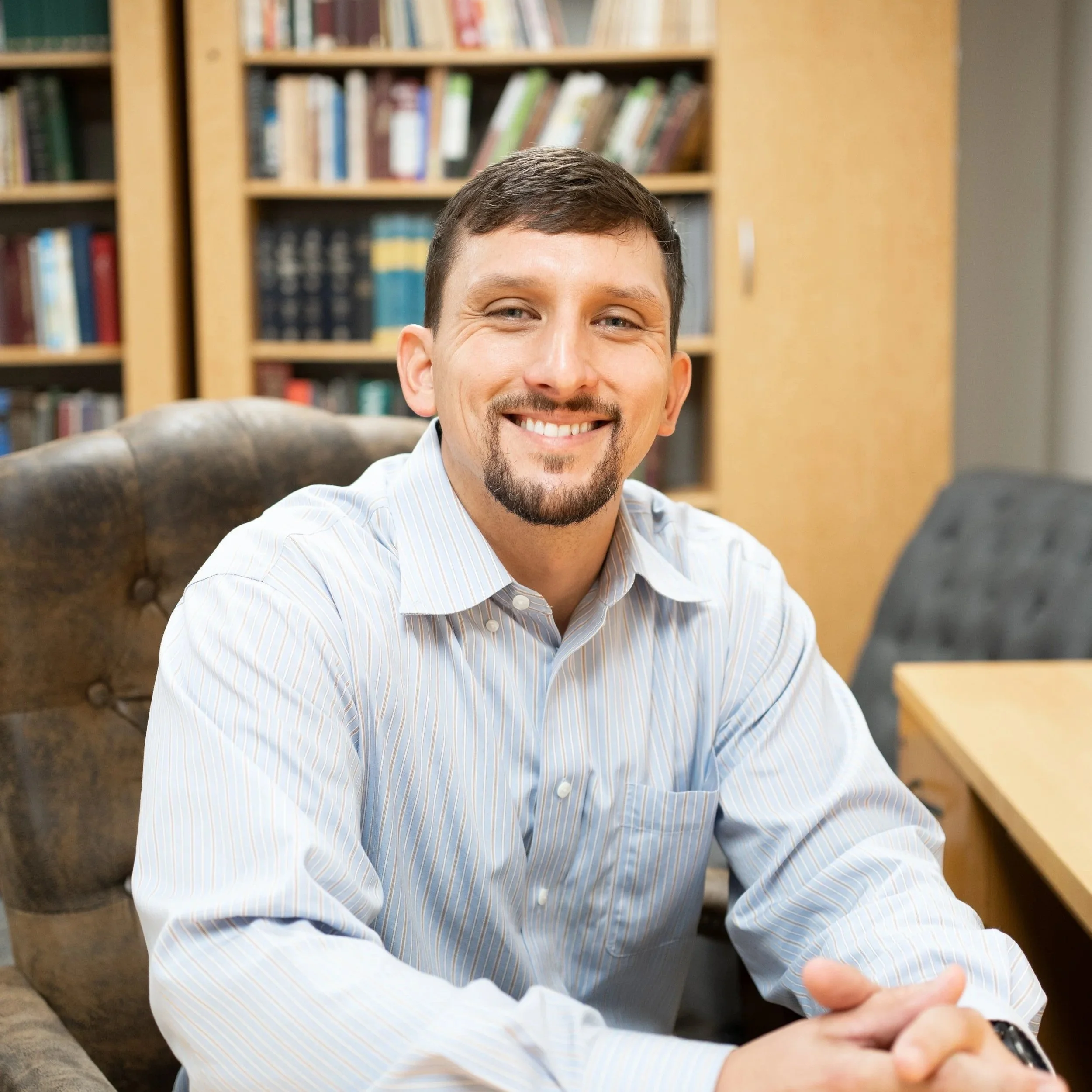 A smiling man with brown hair, a goatee, wearing a light blue striped shirt, sitting in a room with wooden bookshelves filled with books.