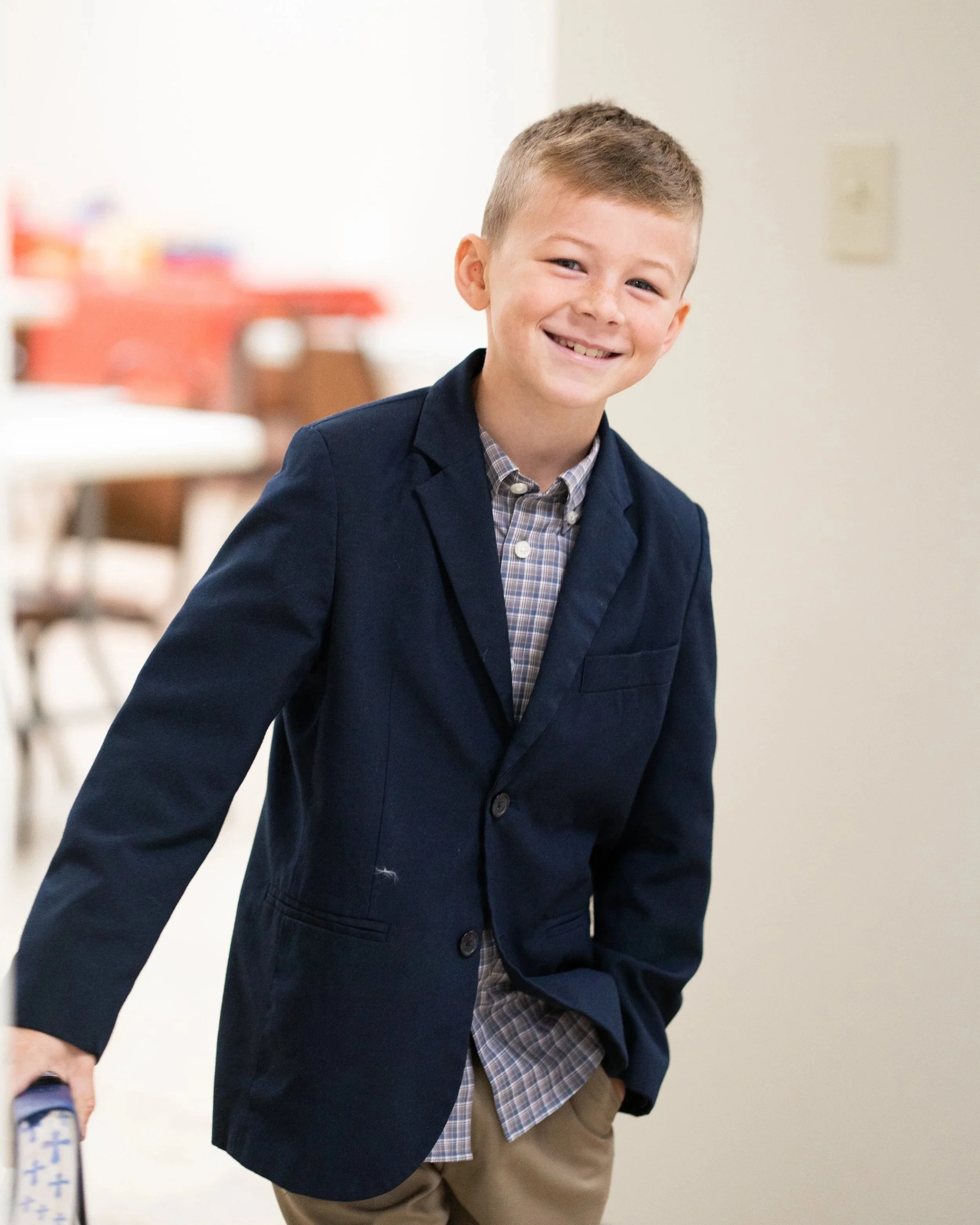 Smiling young boy with short light brown hair, wearing a navy blazer over a checkered shirt, and tan pants, standing indoors.