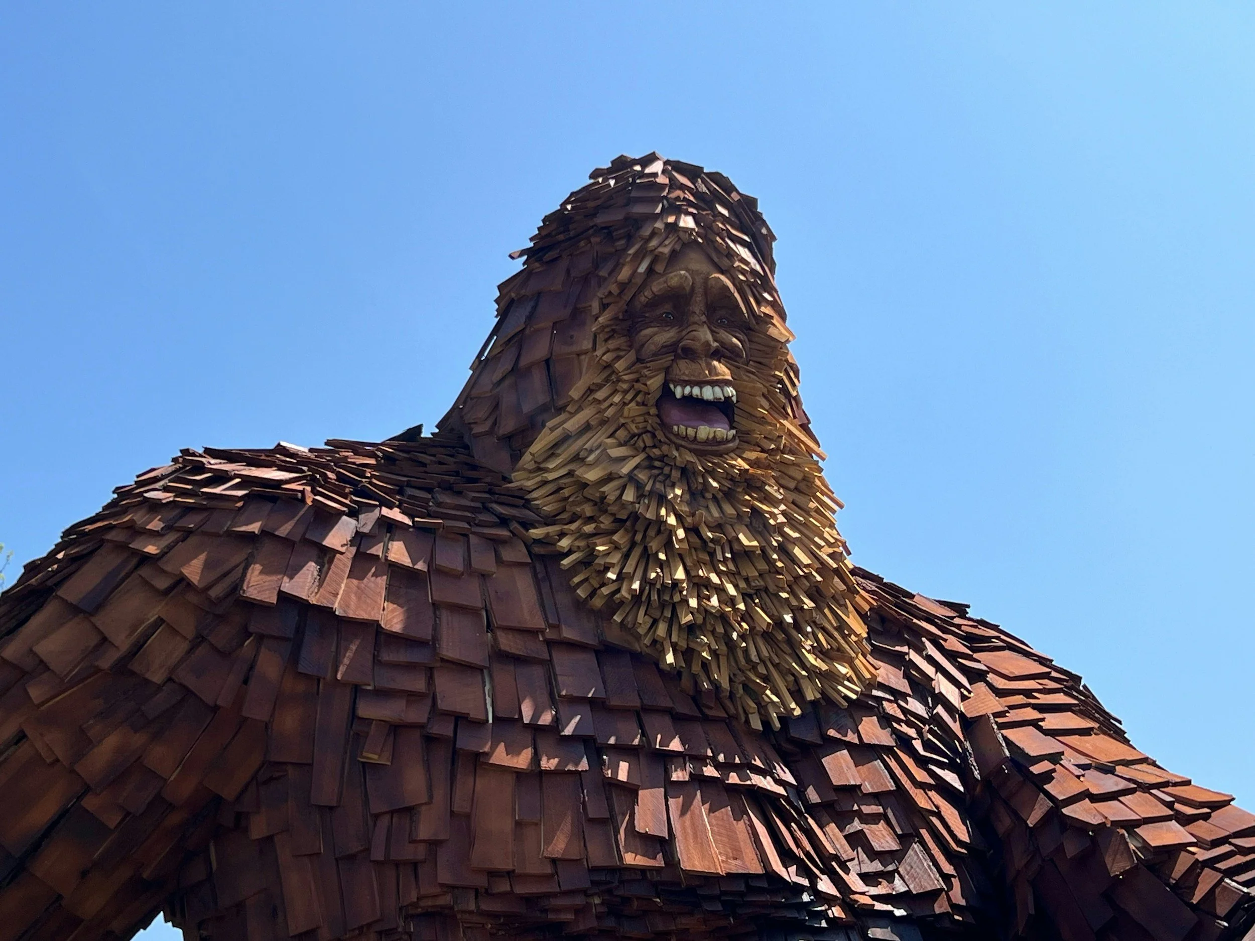 A large wooden sculpture of a mountain gorilla's head and shoulders with an open mouth, made of layered wood pieces against a clear blue sky.