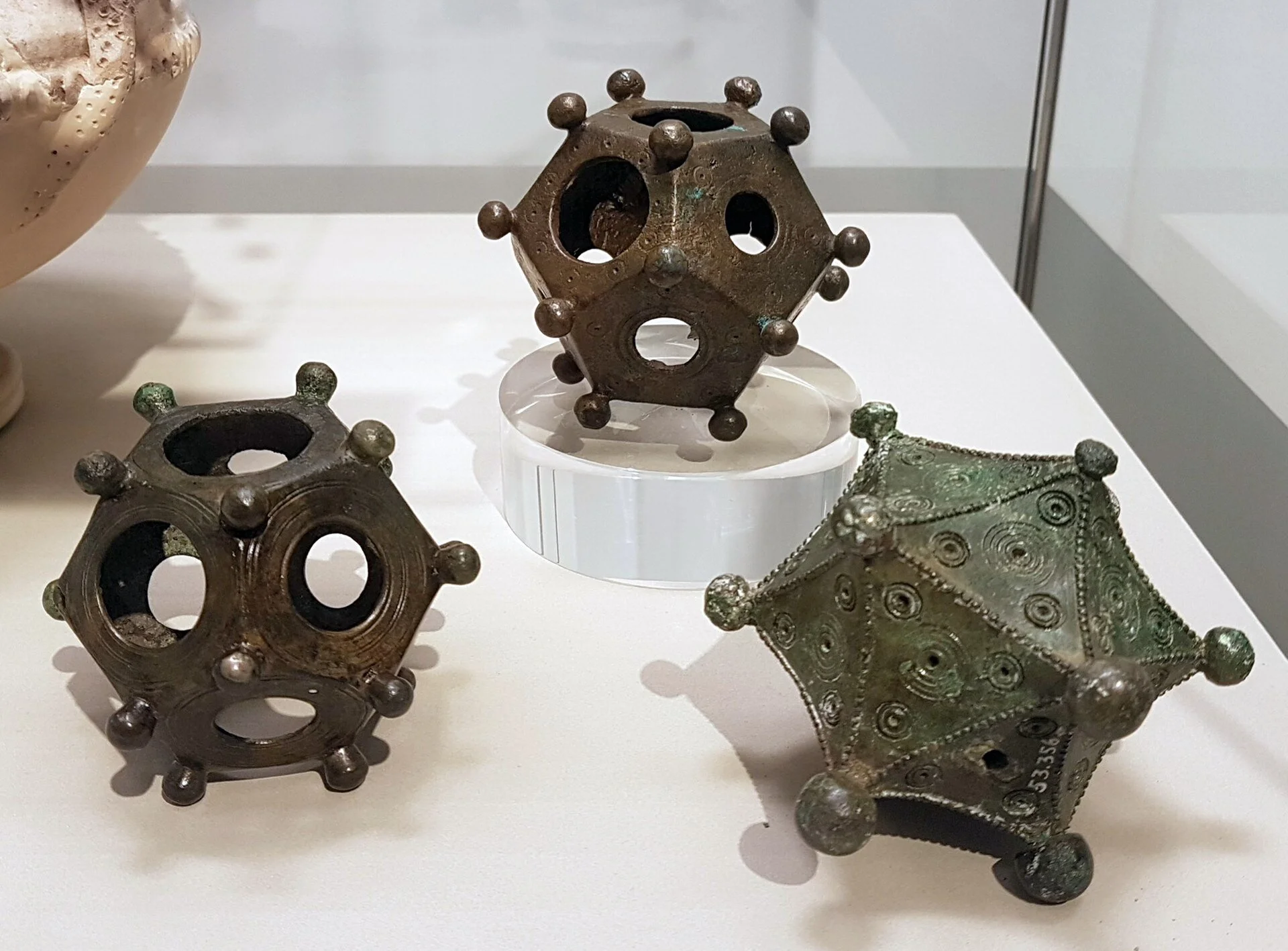 Three ancient metal objects resembling polyhedral shapes with circular holes and small spherical protrusions at the vertices, displayed on a white surface in a museum setting.