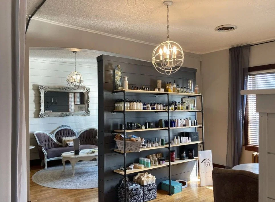 View of a living room with a black shelving unit holding various bottles and containers, a sitting area with two purple velvet tufted sofas, a mirror on the wall, and a modern chandelier hanging from the ceiling.