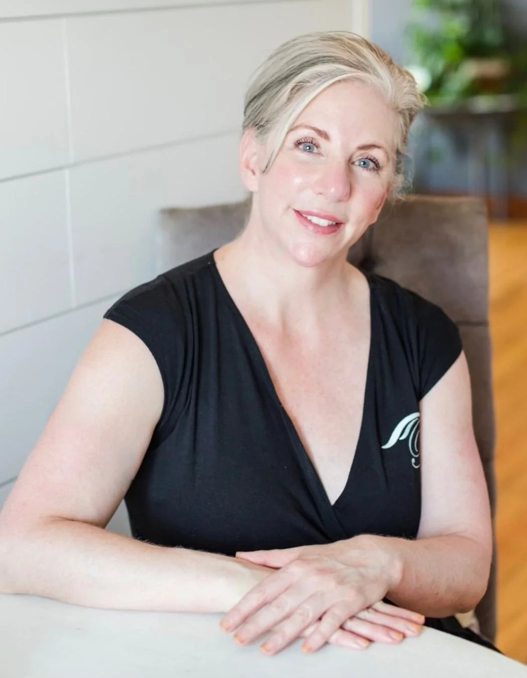 A woman with short gray hair and blue eyes, wearing a black sleeveless top, smiling while sitting at a table in a room with light-colored walls and some greenery in the background.