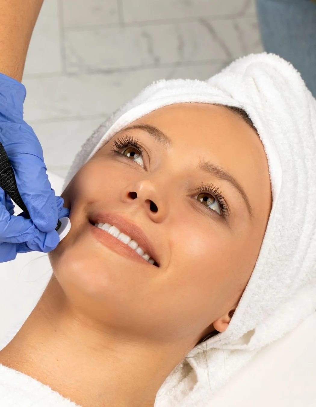 A woman lying down at a spa or clinic, receiving a cosmetic treatment on her face from a professional wearing blue gloves, with her hair covered by a towel.