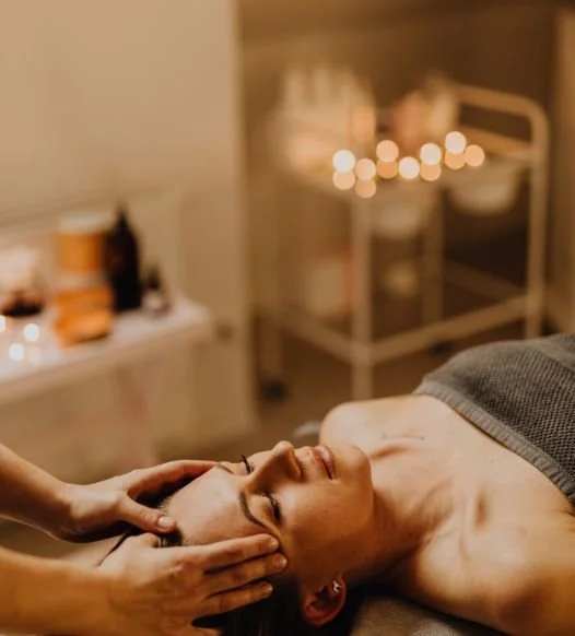 A woman receives a massage with her eyes closed in a spa setting, with candles and skincare products in the background.