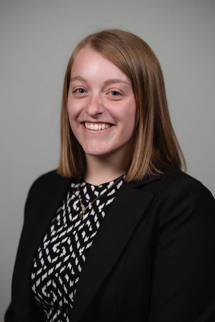 A young woman with shoulder-length light brown hair, smiling, dressed in a black blazer over a black and white patterned top, against a plain gray background.