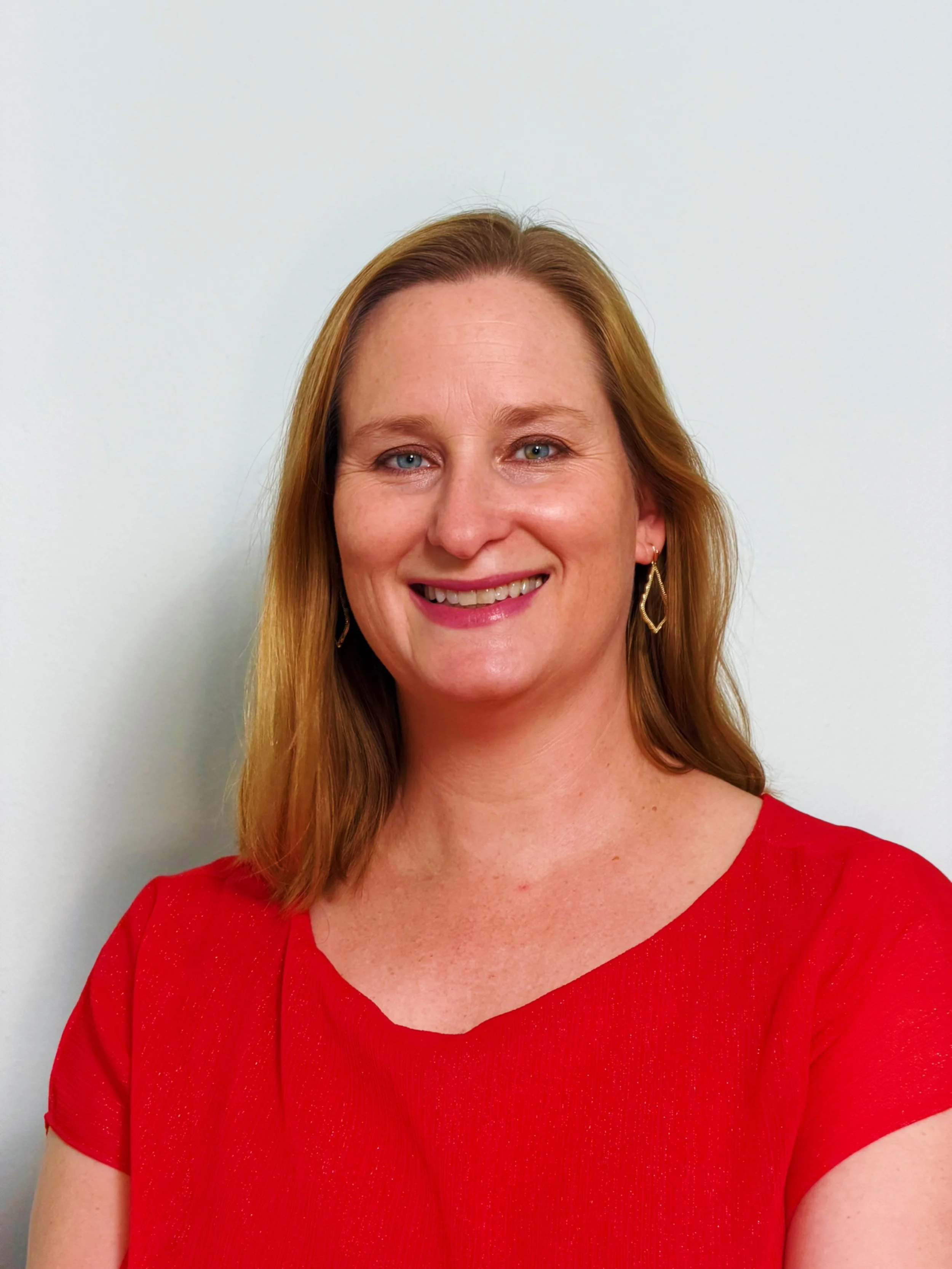 A woman with long red hair wearing a red top, smiling, against a plain white background.