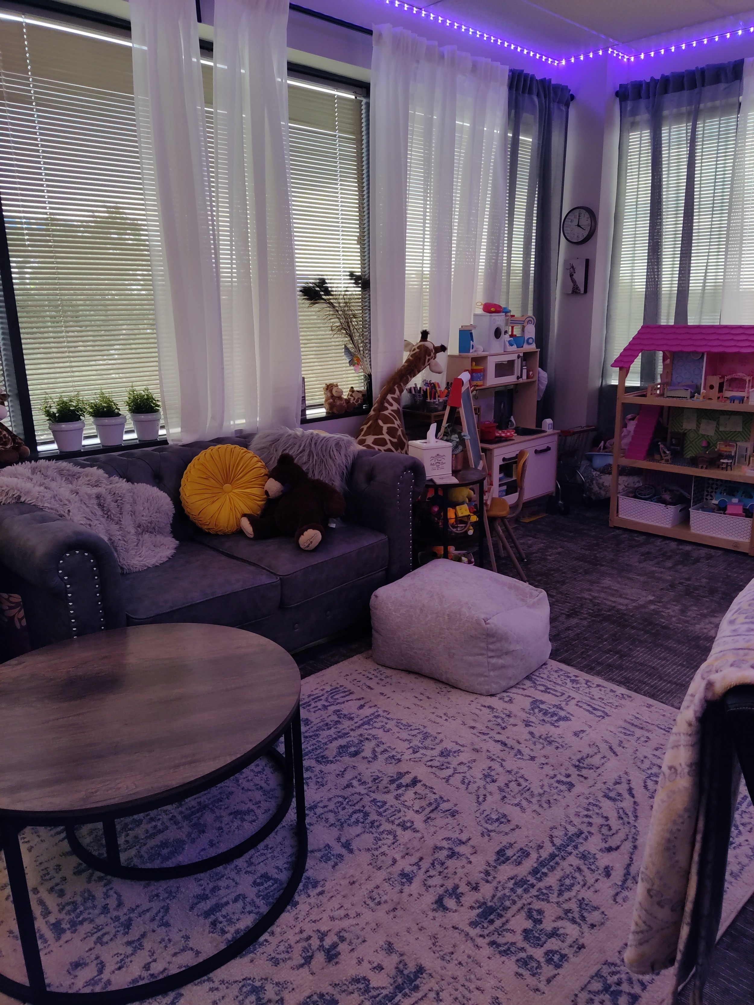 Living room with a large grey couch with stuffed animals and pillows, round wooden coffee table, patterned rug, white curtains, windows with blinds, and purple string lights along the ceiling.
