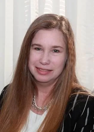 A woman with long red hair smiling in front of white blinds, wearing a black and white striped jacket and a pearl necklace.