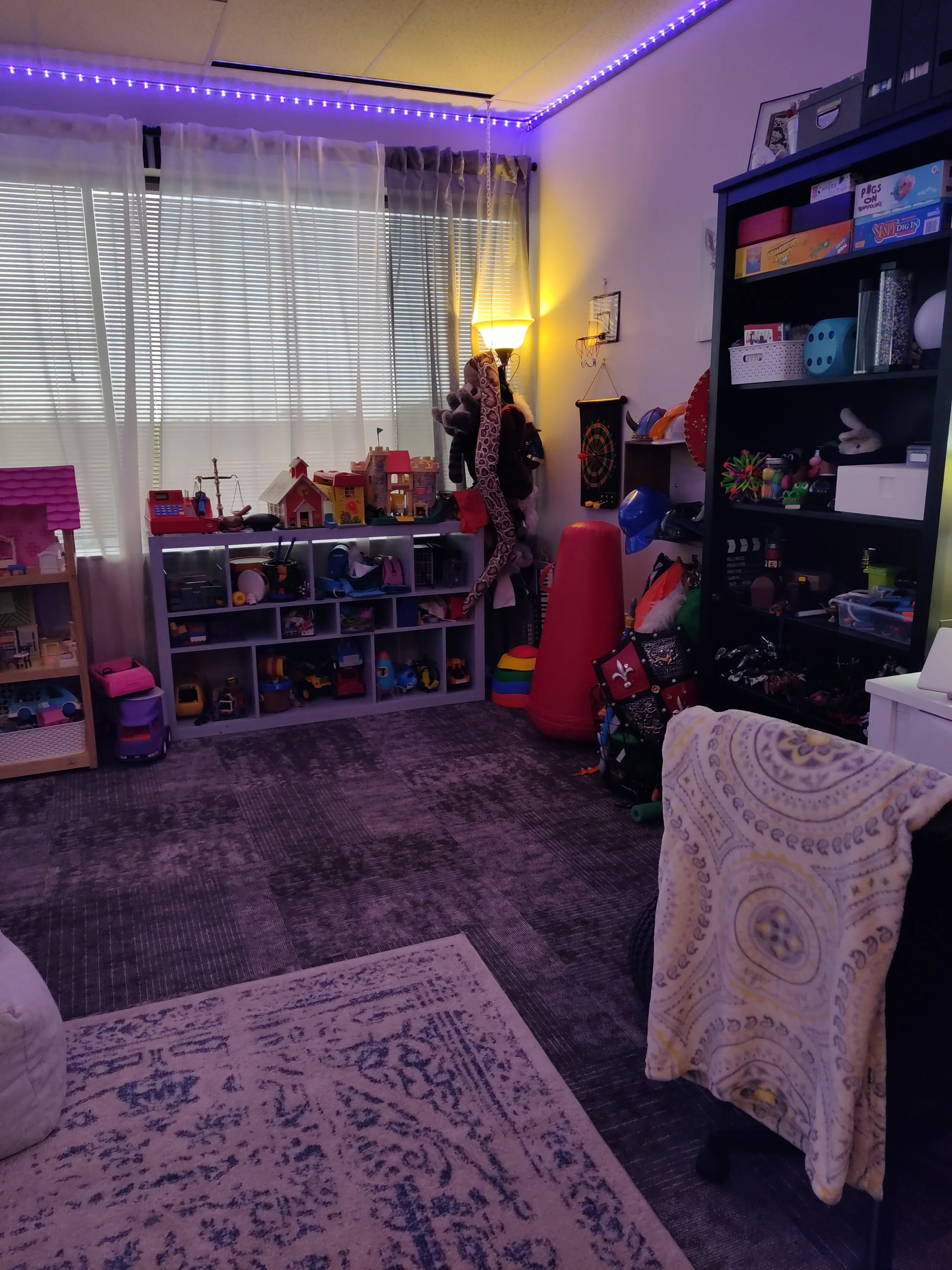 A cozy children's playroom with shelves filled with toys, stuffed animals hanging near a lamp, a workout dartboard on the wall, a large window with curtains, and various toys and play items scattered around the room.