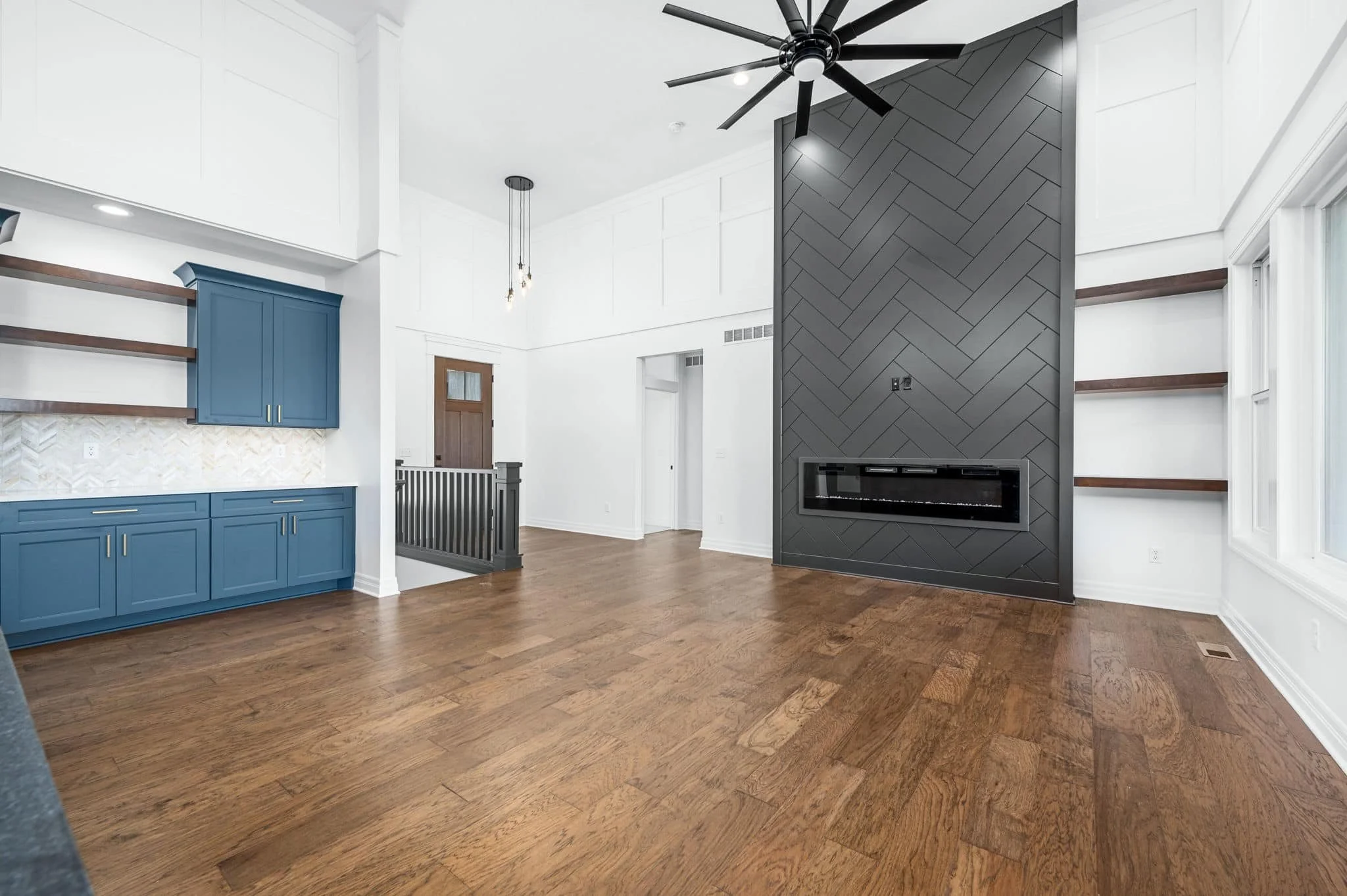 Interior of a modern living room with a black ceiling fan, a dark gray herringbone wood fireplace wall, white walls, a built-in blue cabinet with gold handles, and wooden flooring.