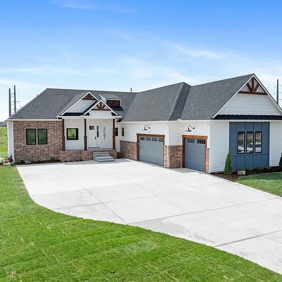 Front view of a modern house with a concrete driveway, brick and white siding exterior, and a gabled roof under a partly cloudy sky.