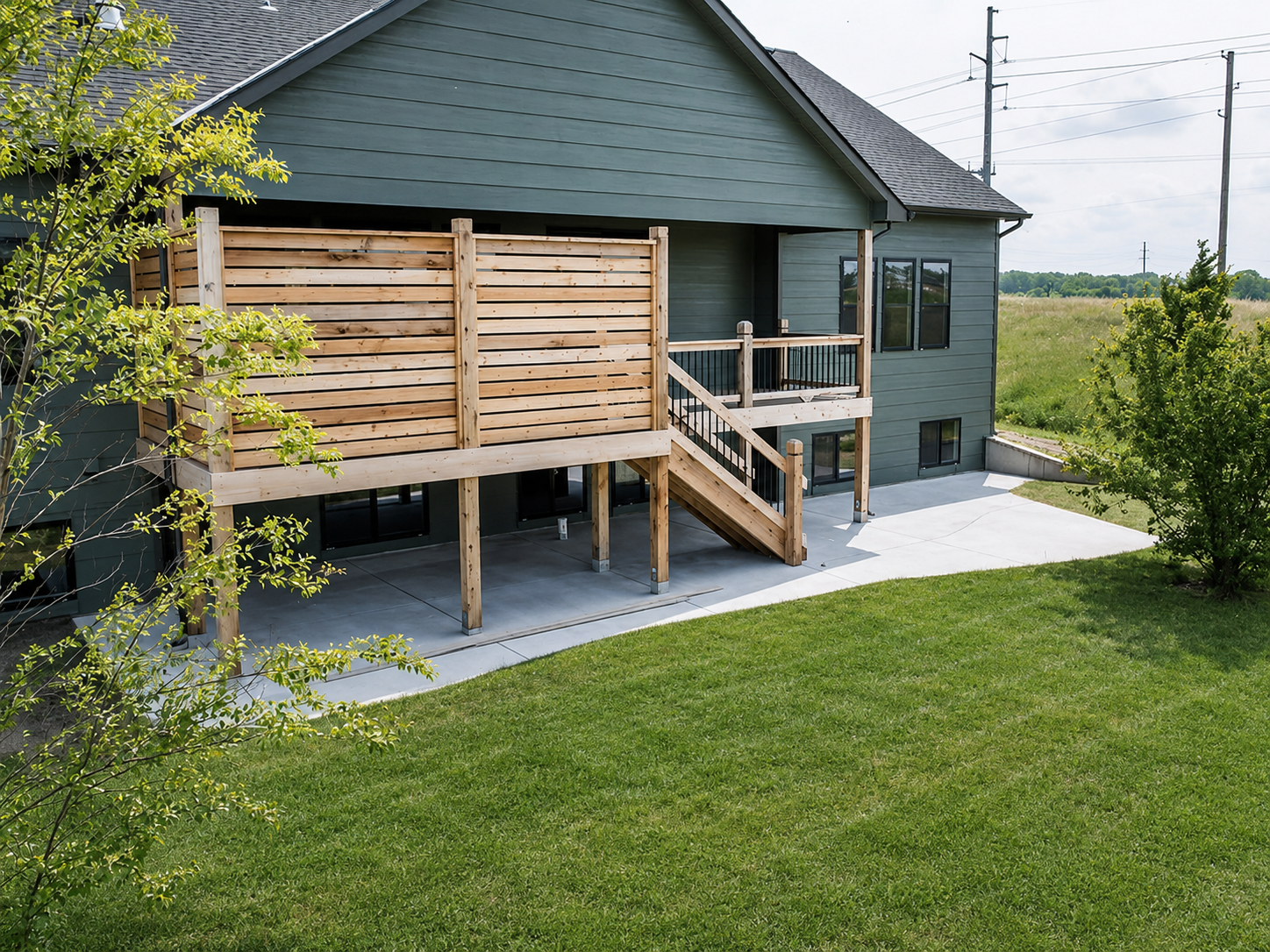 Backyard view of a modern house with a wooden deck and stairs, green grass lawn, and trees, with power lines in the background.