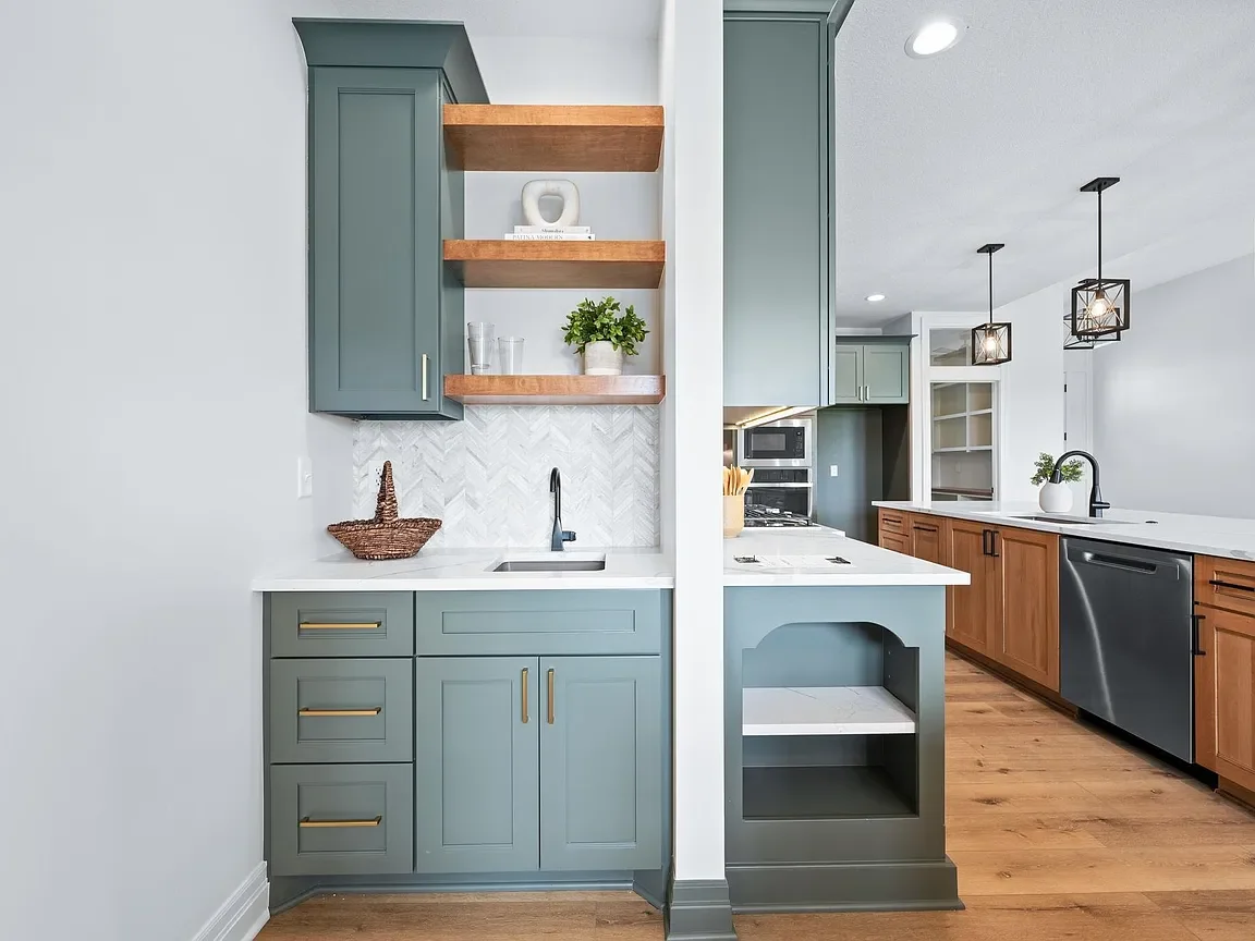 A modern kitchen with green and wood cabinetry, open shelves, a black faucet, a white marble backsplash, and a wood floor.