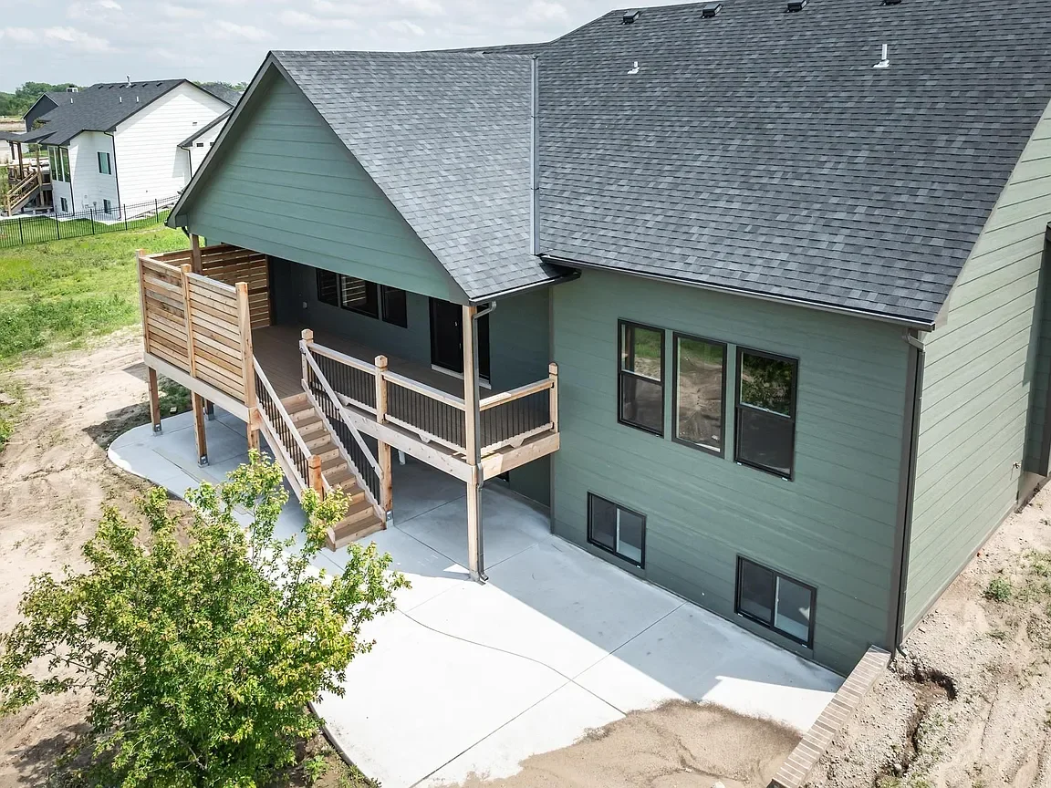 A newly constructed green house with a dark gray roof and a wooden deck, on a concrete patio, with a staircase leading up to the deck and surrounding grassy yard.