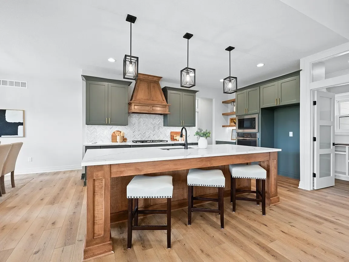 Modern kitchen with a large island, gray cabinets, a wooden range hood, pendant lighting, and wooden flooring.