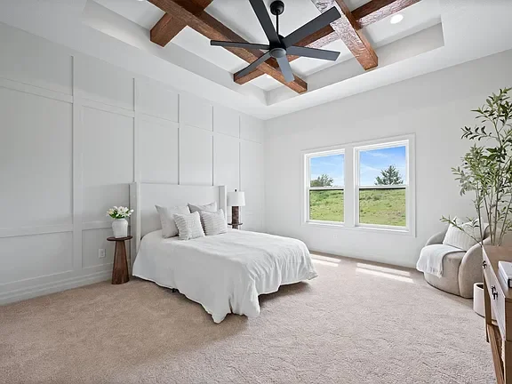 Bedroom with white walls, a bed with white linens, a nightstand with a lamp, large windows showing a green landscape, and a ceiling fan with wooden beams.
