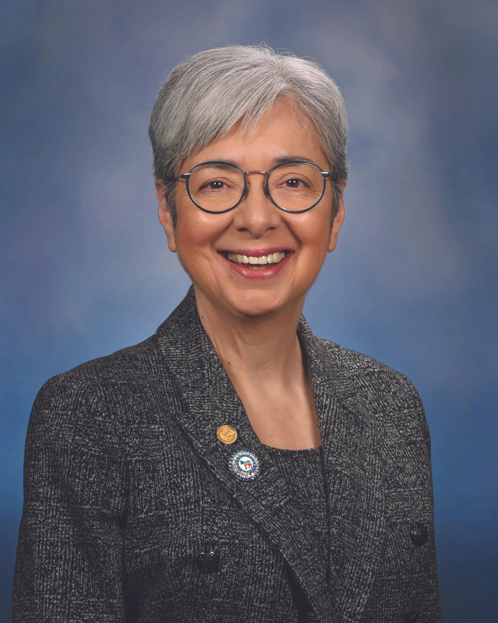 Close-up portrait of an elderly woman with short gray hair, wearing glasses and a black textured blazer, smiling against a blue background with cloud patterns.