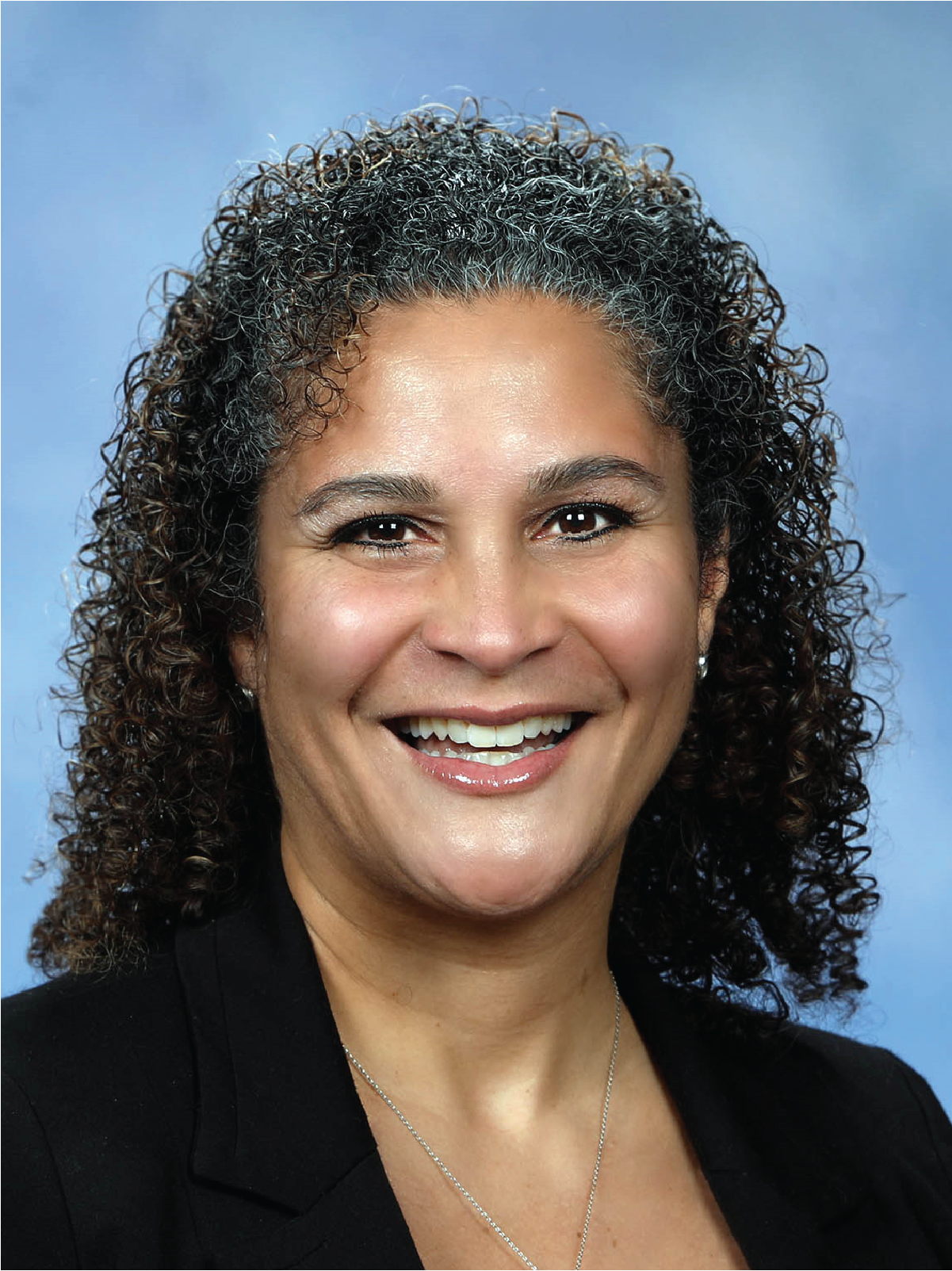 A middle-aged woman with dark, curly hair graying at the temples, smiling against a light blue background, wearing a black blazer and a delicate silver necklace.
