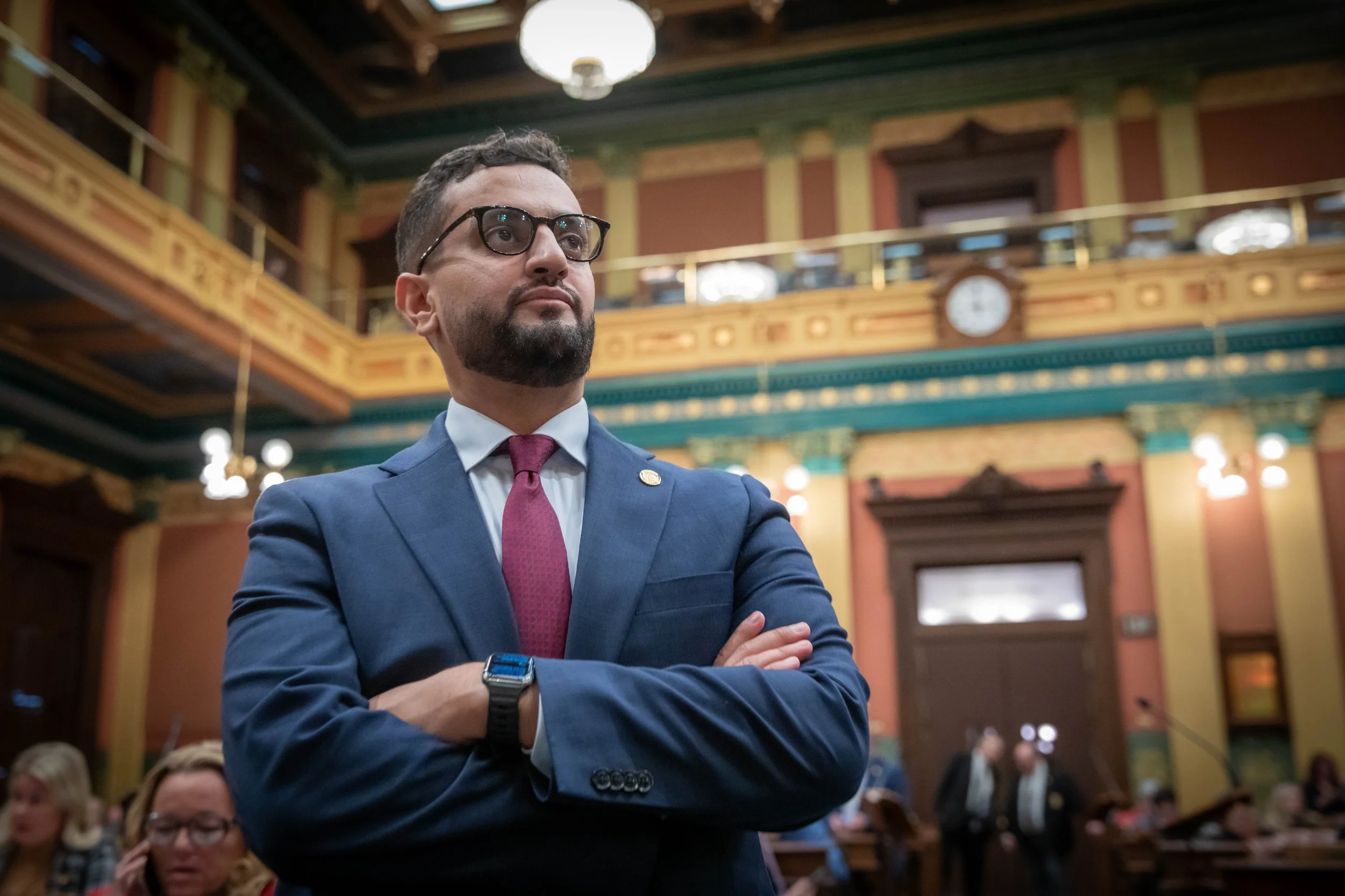 A man wearing glasses and a blue suit with a red tie, standing with crossed arms in a historic building with ornate woodwork and a clock in the background. There are people sitting around, suggesting a formal event or gathering.