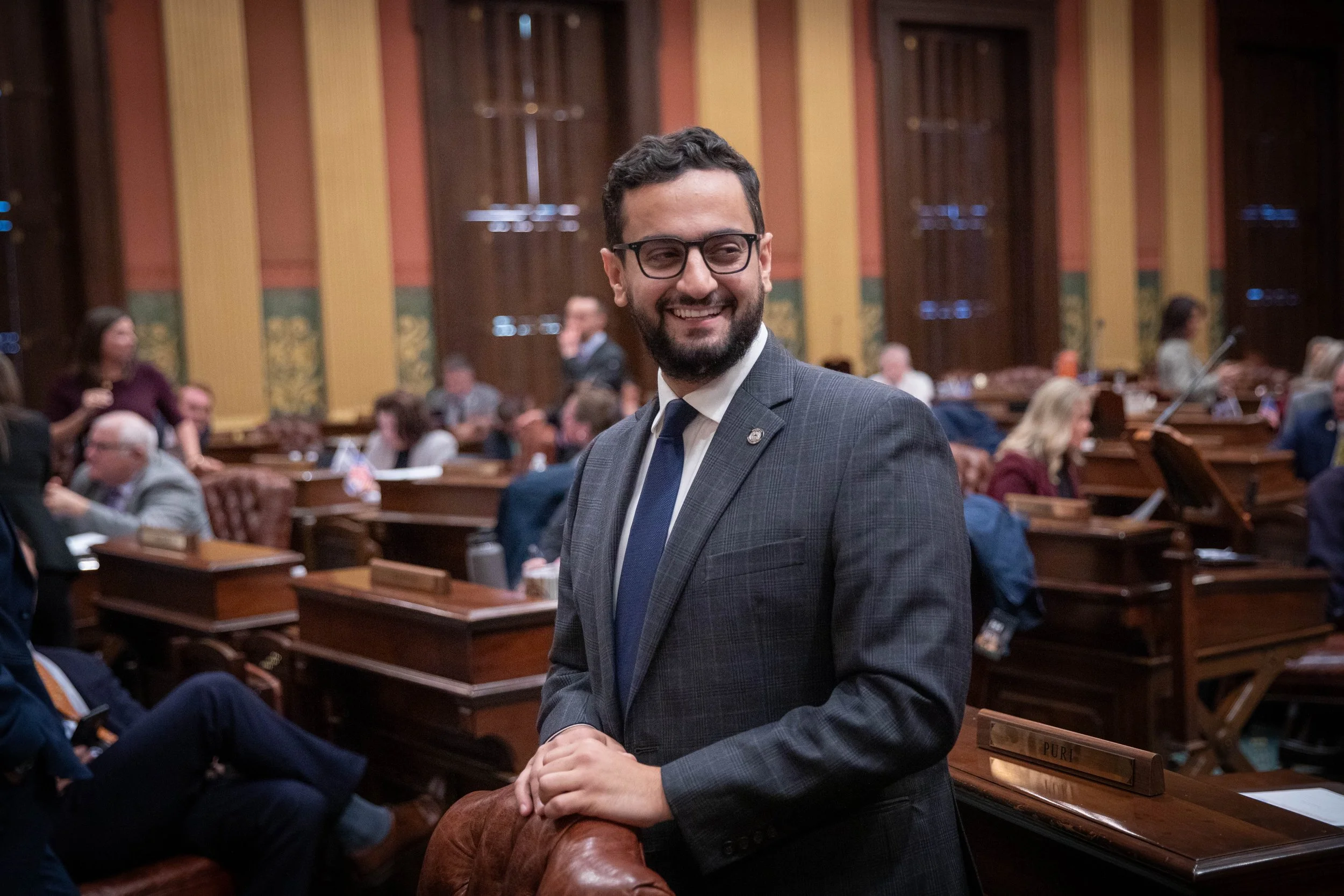 A smiling man with glasses and a beard wearing a suit and tie, standing in a large, wood-paneled legislative chamber with people seated at desks in the background.