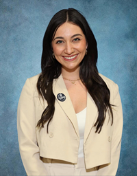 A woman with long dark hair wearing a light-colored blazer and smiling against a blue background.