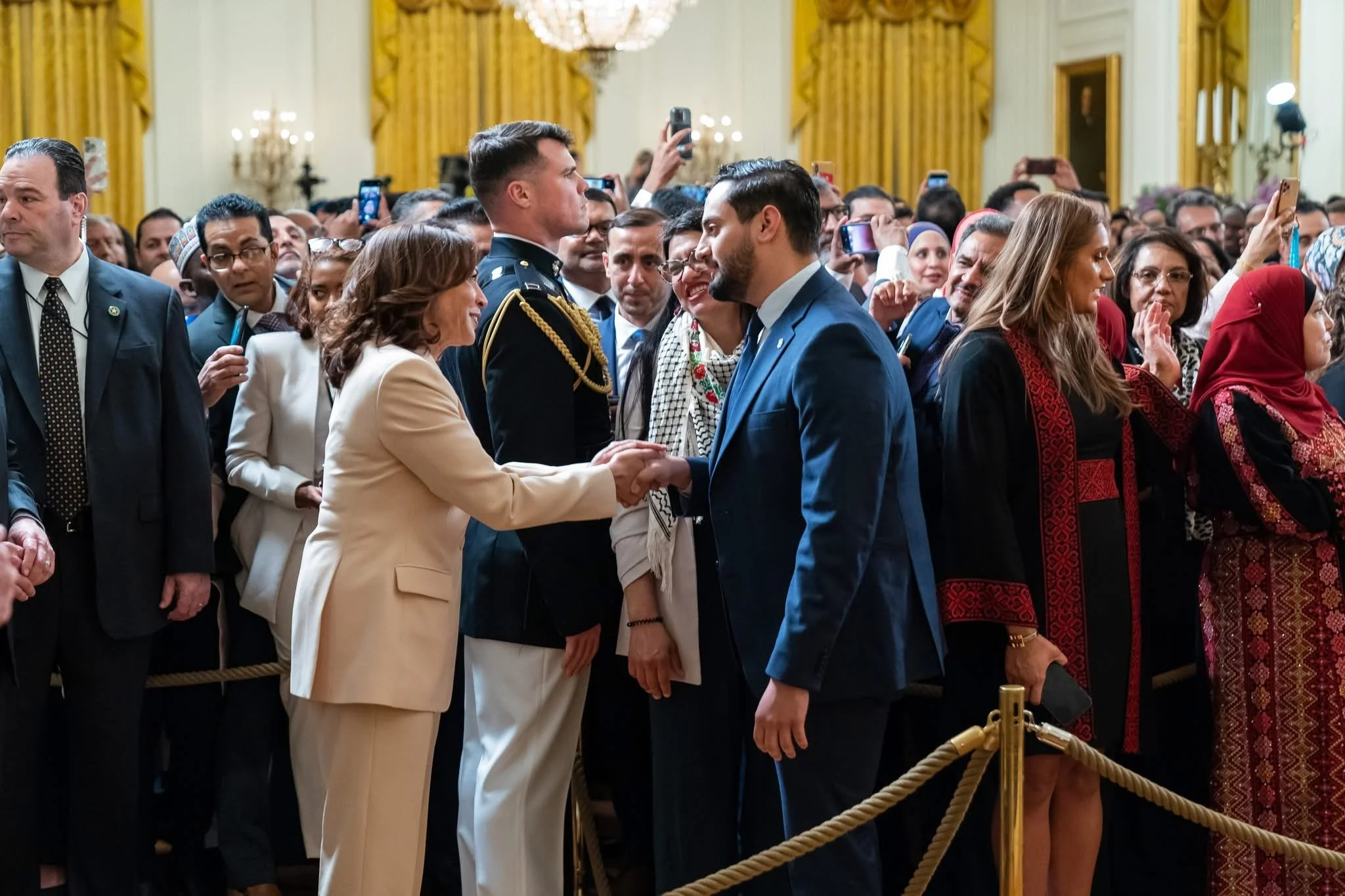 A woman in a beige suit shaking hands with a man in a blue suit among a crowd of people in a formal setting.