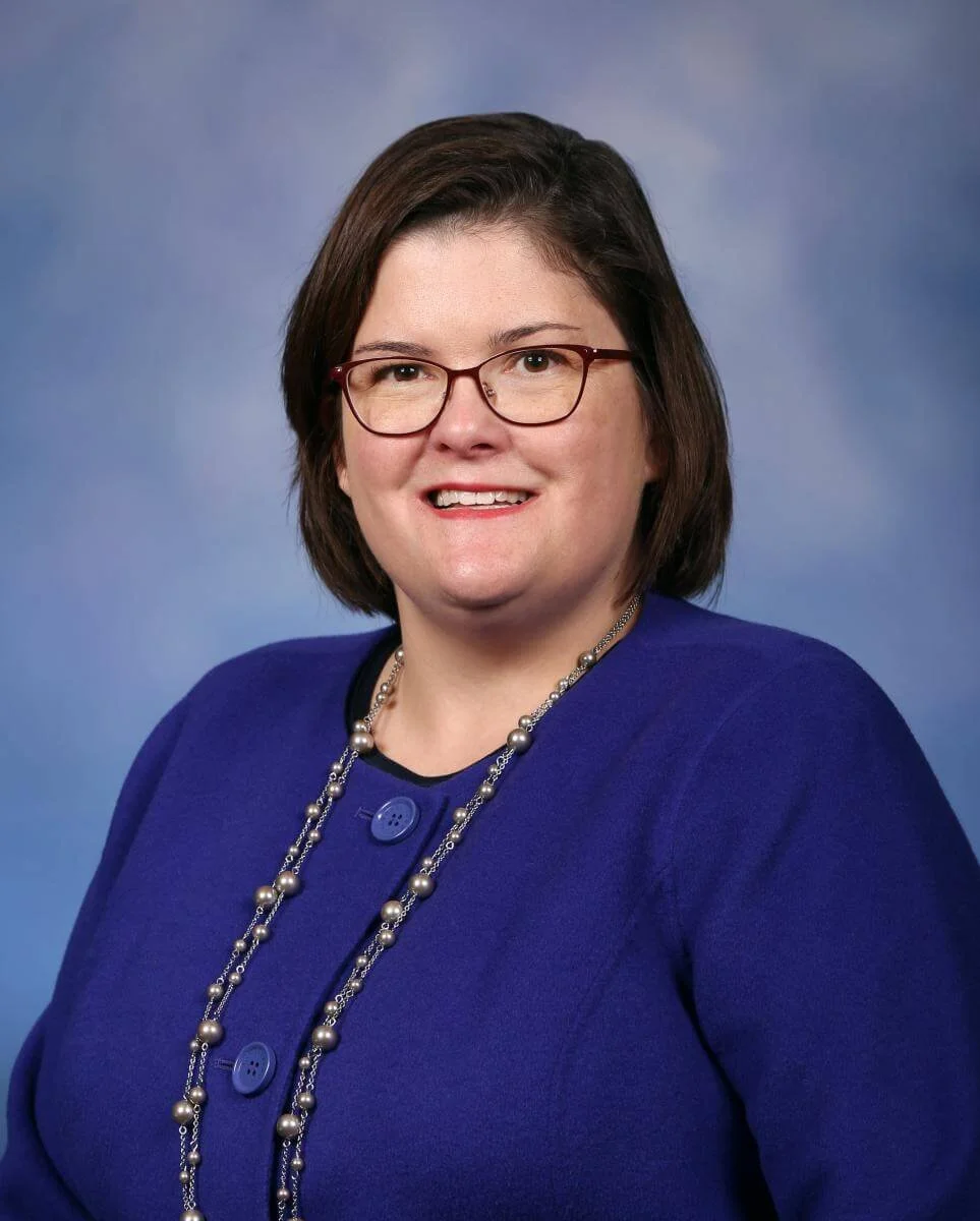 A woman with brown hair and glasses wearing a blue blazer and a pearl necklace against a blue background.