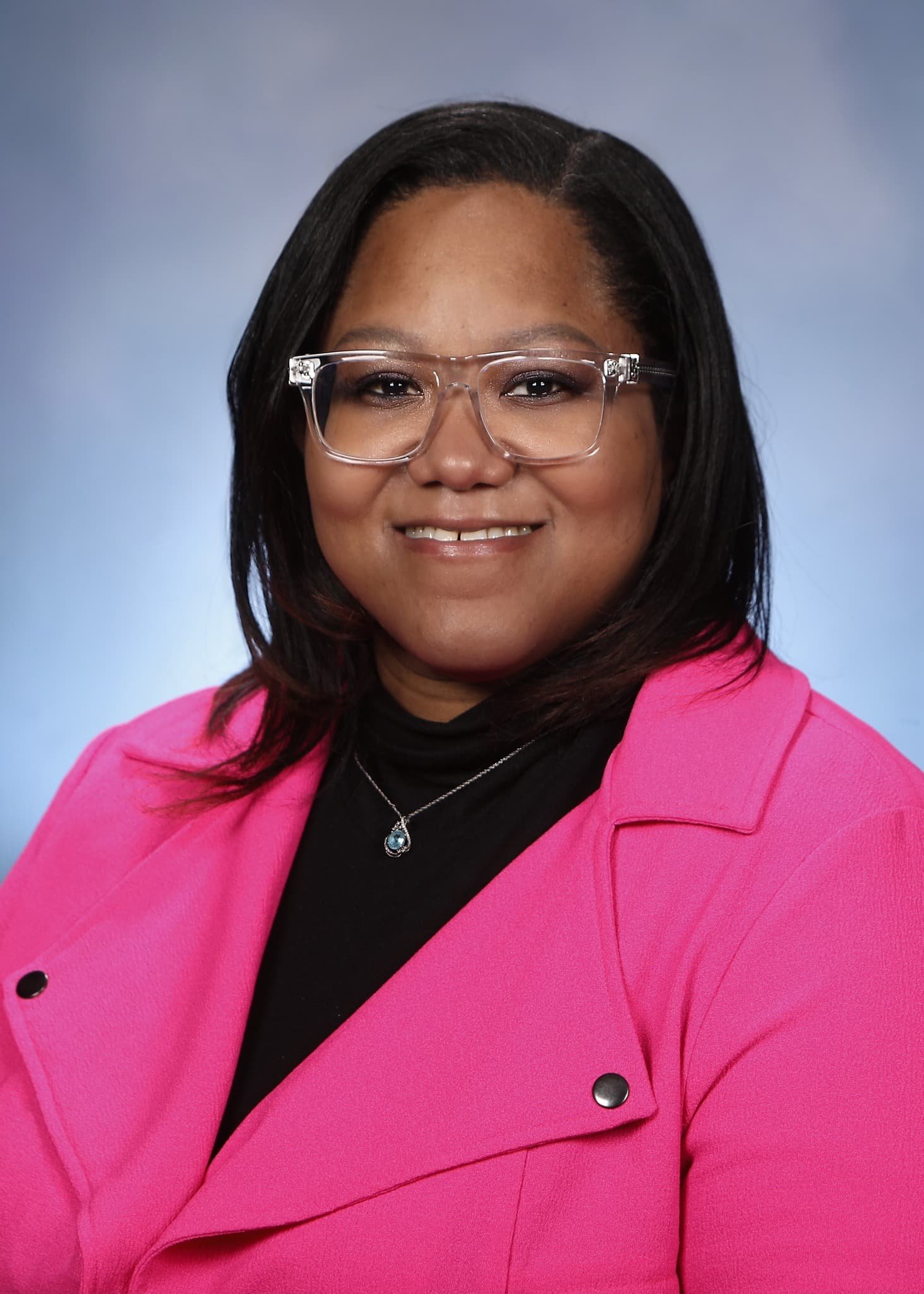 A woman with shoulder-length black hair wearing clear glasses, a bright pink blazer, and a black top. She has a necklace with a blue gemstone pendant and is smiling against a neutral blue background.