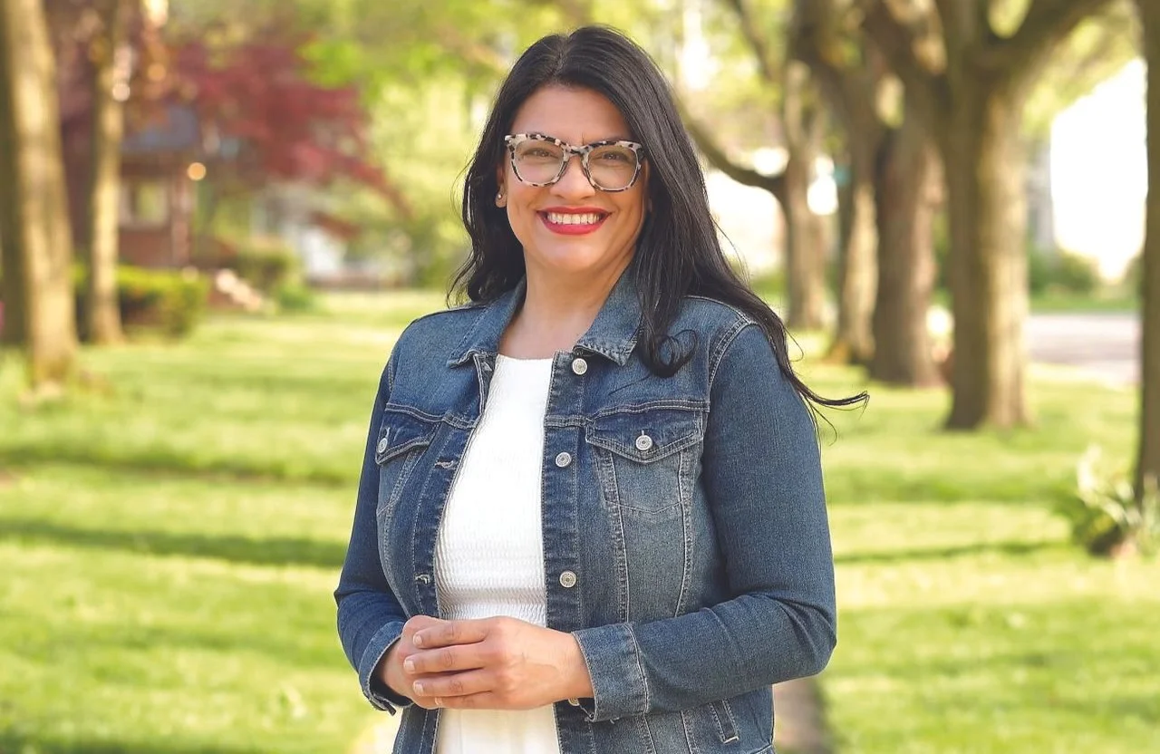 A woman with dark hair, wearing glasses, a white top, and a denim jacket, smiling with hands clasped together outdoors in a park-like setting with trees and green grass.