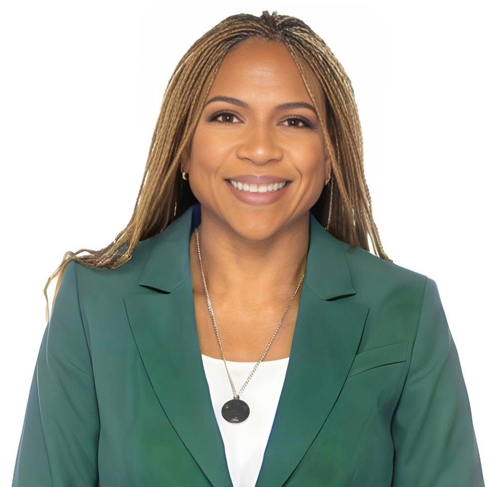 A smiling woman with long blonde hair wearing a green blazer, white top, and a necklace, against a white background.