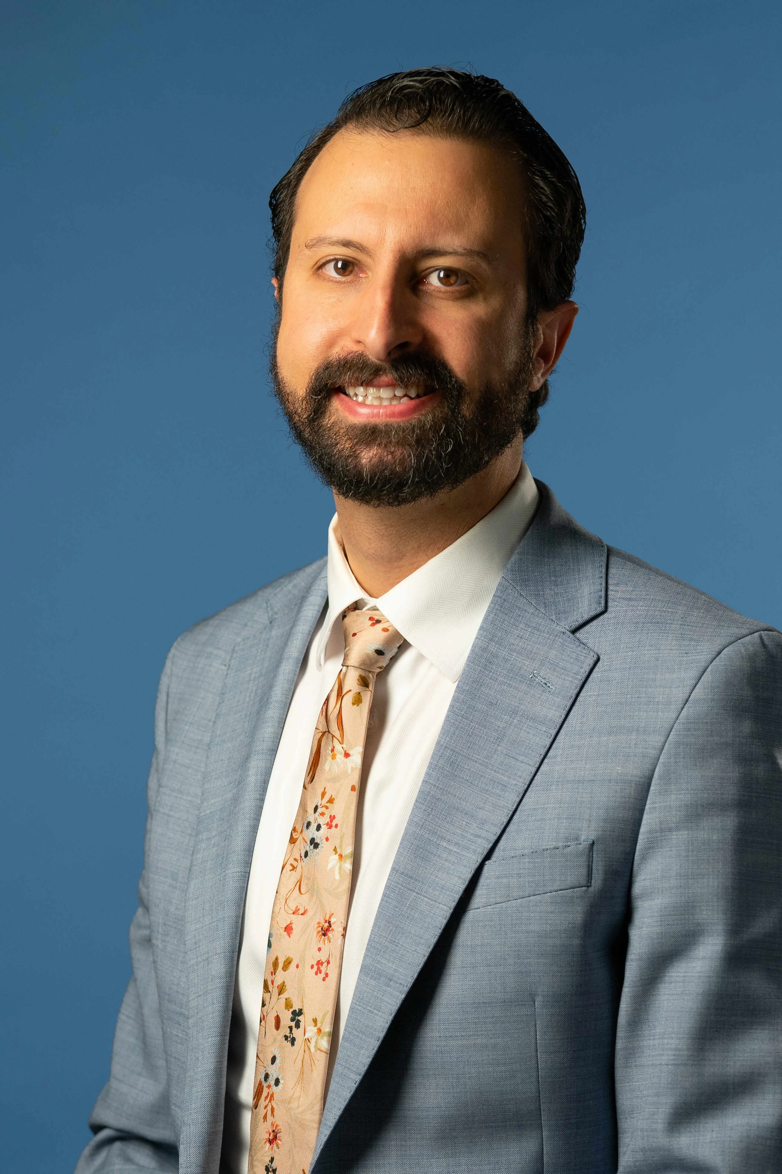 Headshot of a man with dark hair, beard, and mustache wearing a light blue suit, white shirt, and a floral tie, standing against a blue background.