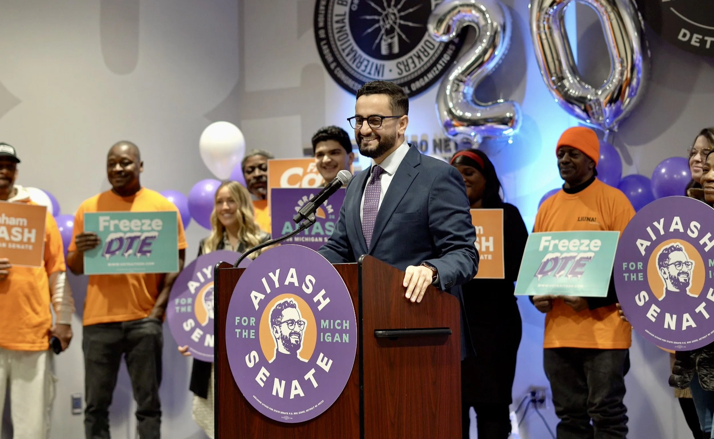 A group of people at a political event, with a man in a suit speaking at a podium decorated with a campaign logo for Michigan Senate. Behind him are individuals holding signs with campaign slogans and wearing orange shirts, with some wearing orange hats. Large balloons and a circular emblem are seen in the background.