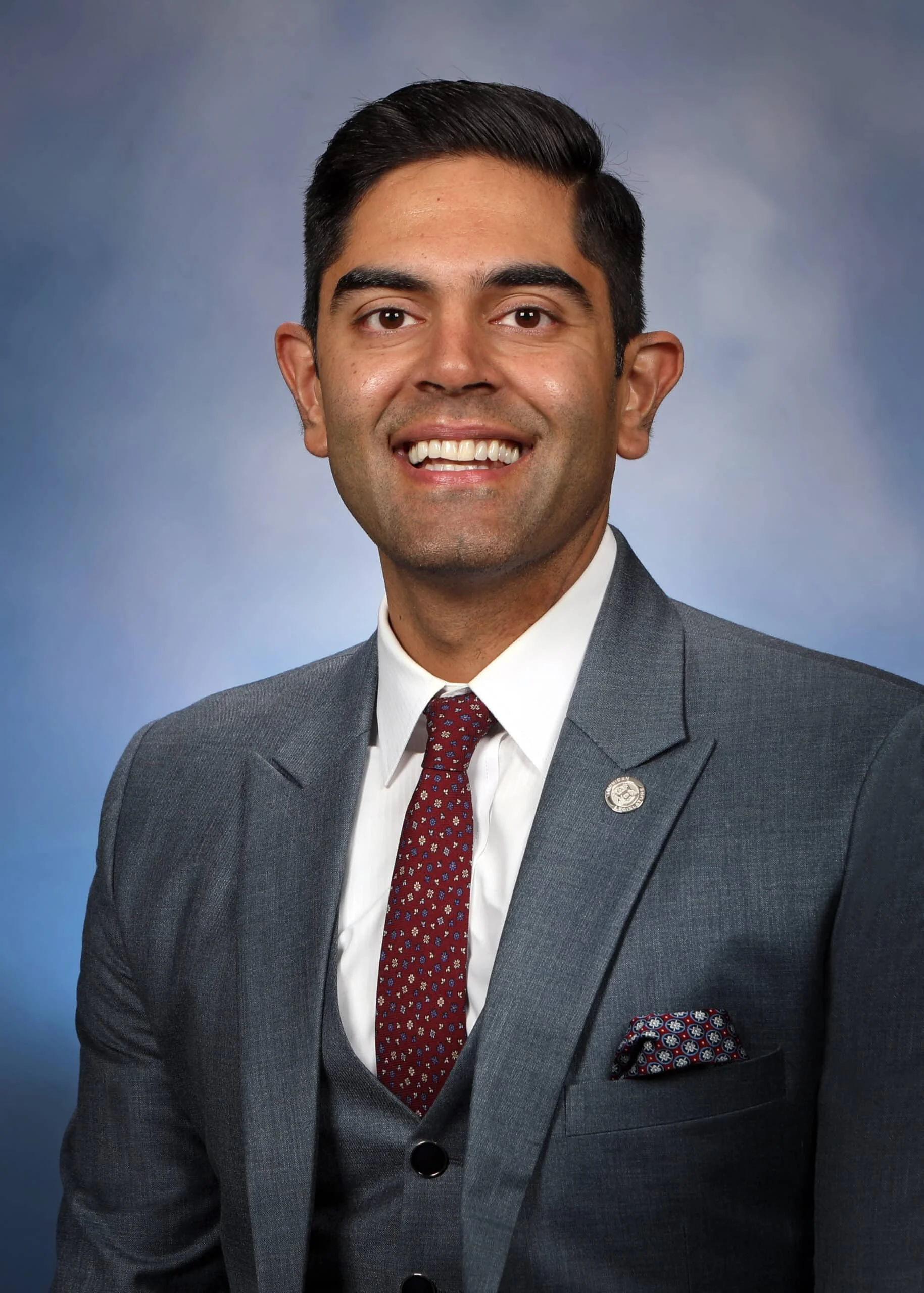 Portrait of a smiling man with dark hair, wearing a gray suit, white shirt, and a red patterned tie, against a blue gradient background.