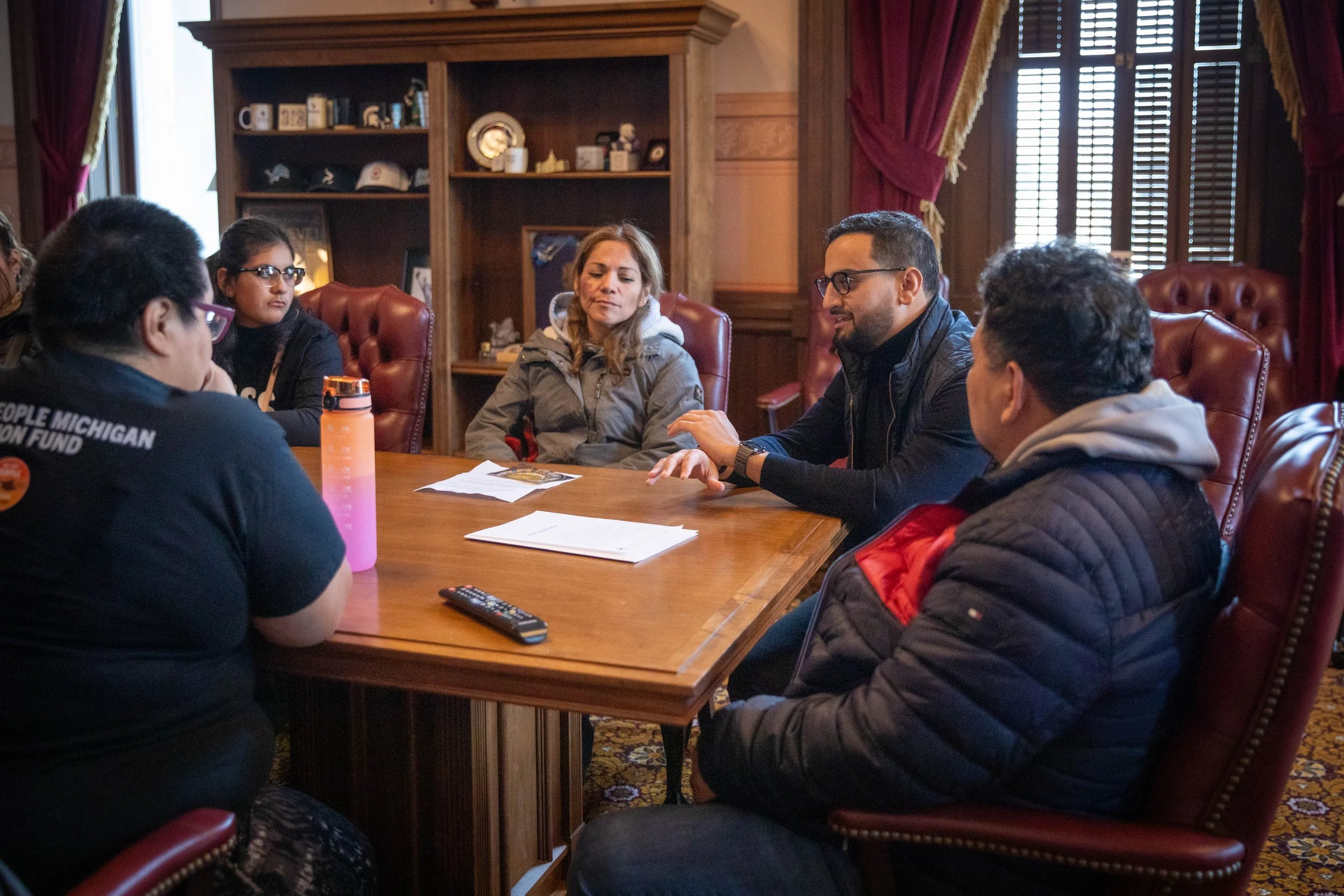 A group of people sitting around a wooden table in a room with wood-paneled walls and large windows. Some are facing each other, engaged in conversation, with papers and a remote control on the table, and a bookshelf with decorative items in the background.