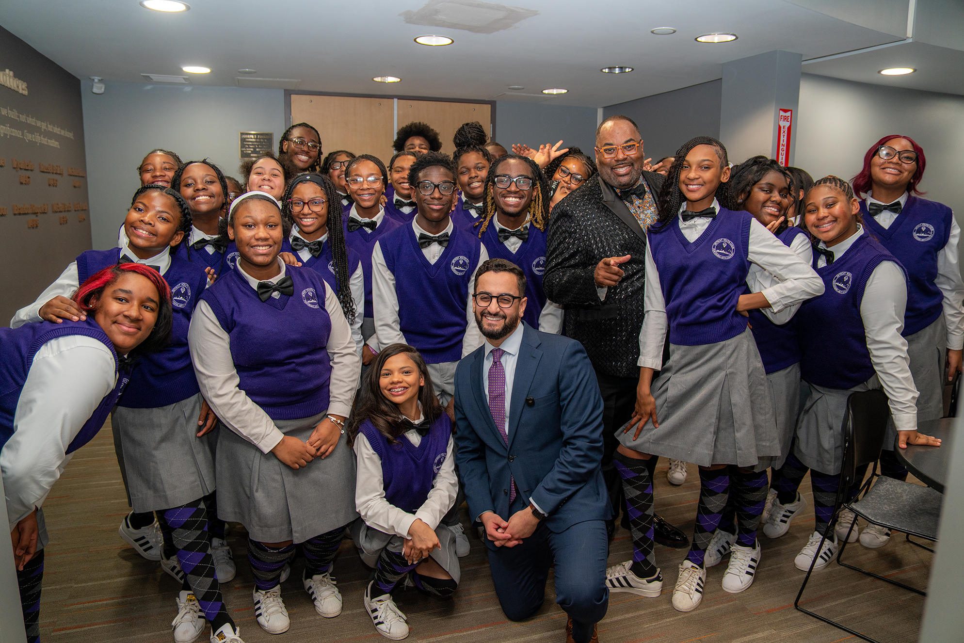A group of students in purple and gray uniforms posing with two men in formal attire in a classroom or auditorium setting.
