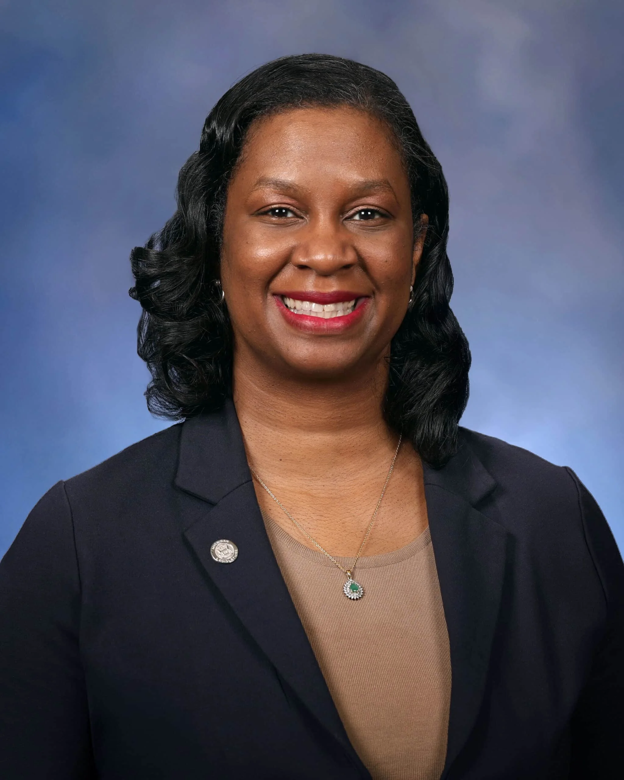 Headshot of an African American woman with black wavy hair and a warm smile, wearing a dark blazer, beige top, and a necklace with a green stone pendant, dressed professionally against a blue gradient background.