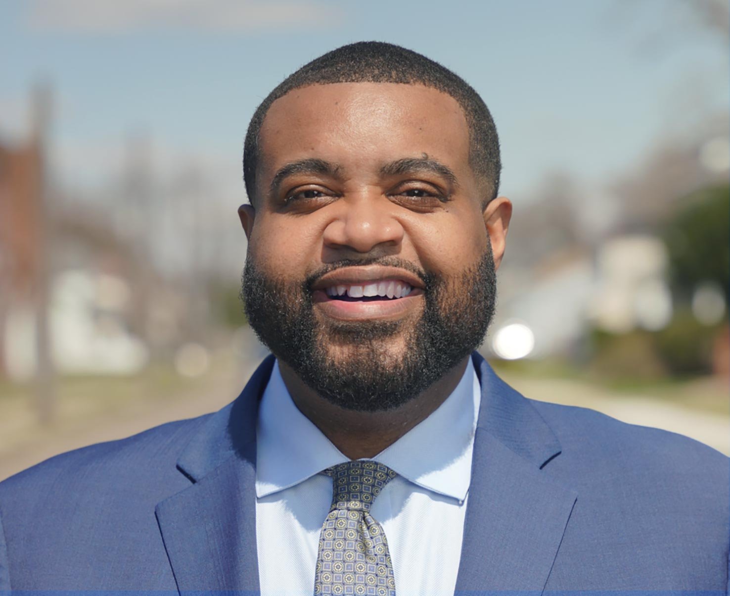 A smiling man in a blue suit, white shirt, and patterned tie outdoors with a blurred background of trees and buildings.