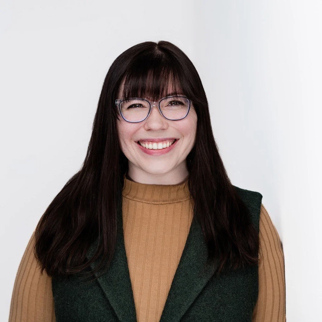 A woman with long dark hair wearing glasses, a tan turtleneck, and a dark vest, smiling against a plain white background.