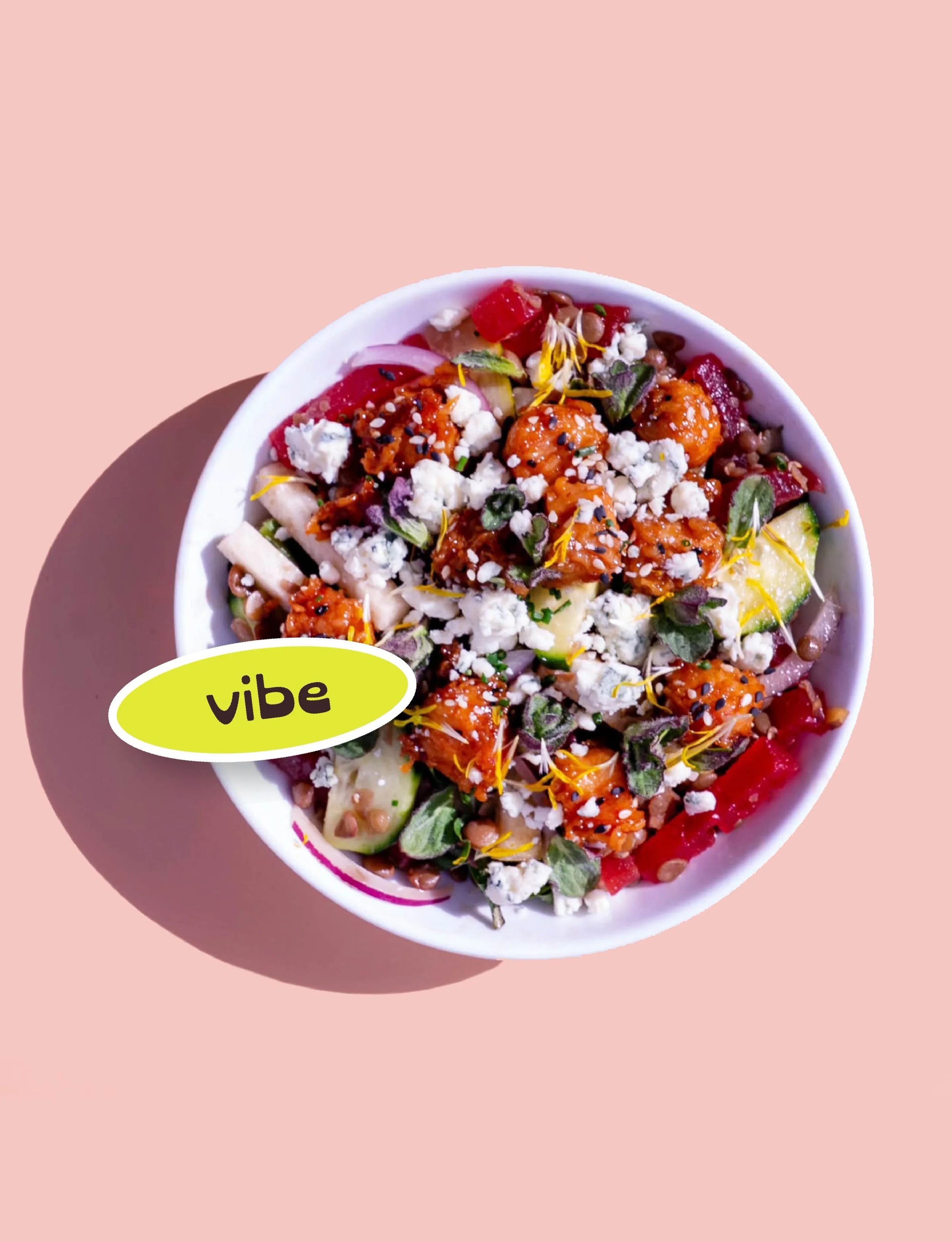 A bowl of colorful salad with vegetables, cheese, and toppings, on a pink background with a yellow label that says 'vibe'.