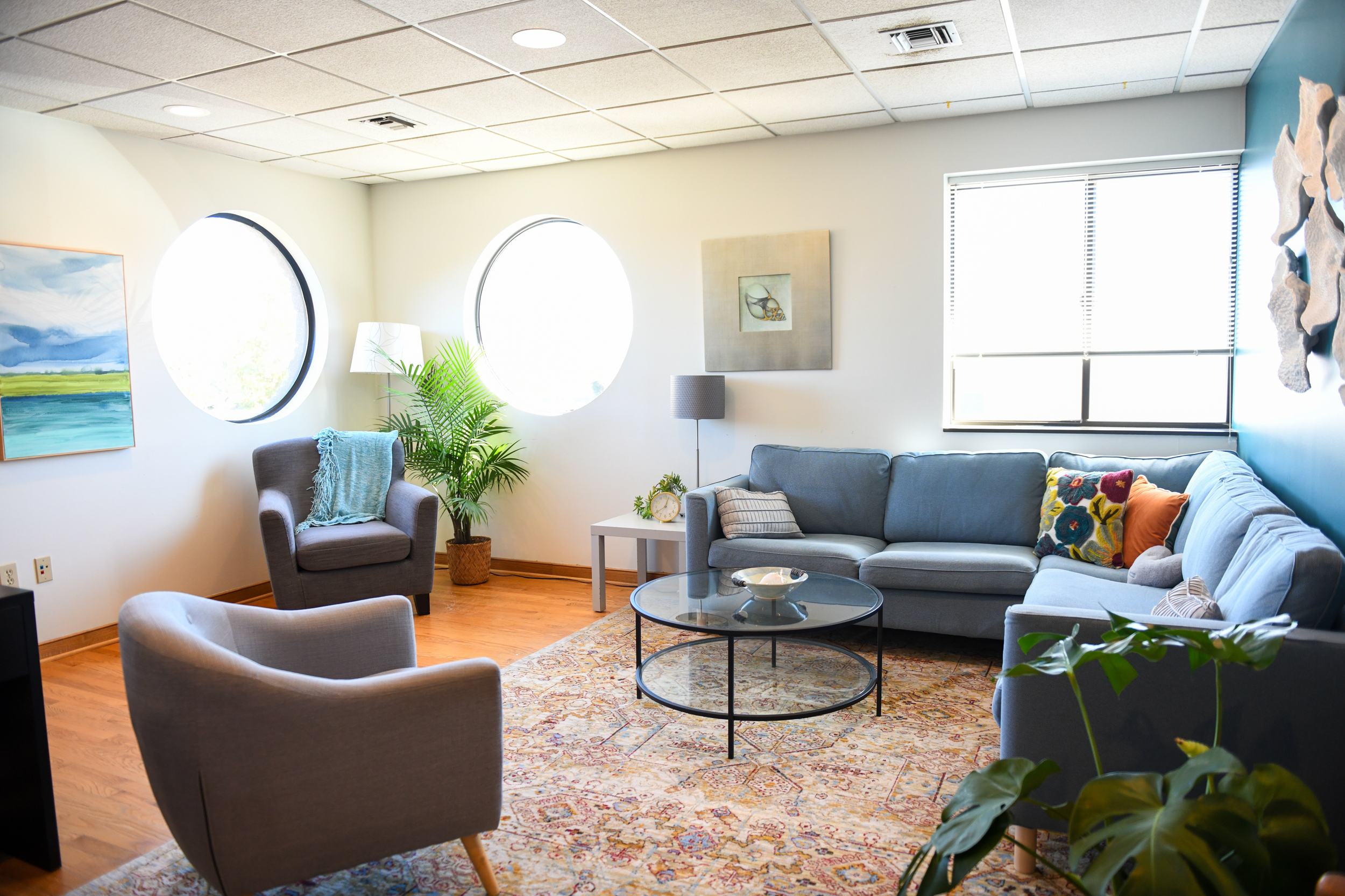 Interior of the Watermark office located in Norfolk, VA. The room features a gray sofa and armchair with blue decorative blankets and pillows. Bright light seeps from the circular and rectangular windows, which creates a calming environment.