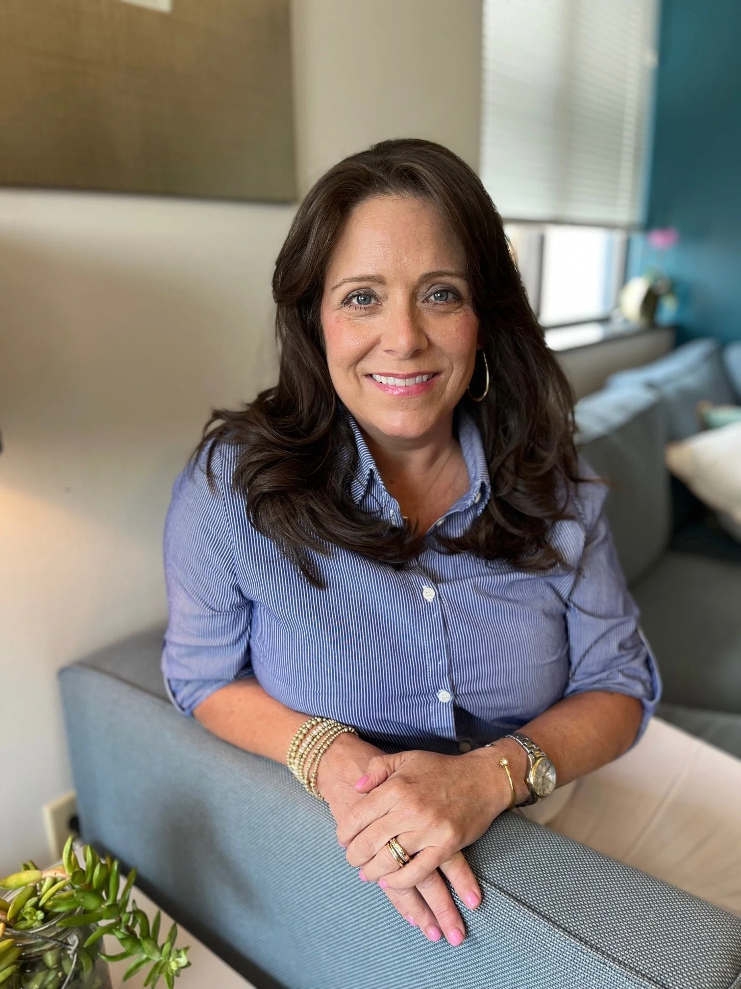 A woman with long dark hair, wearing a light blue striped shirt, is sitting on a gray upholstered chair in a room with a window and a blue wall. She is smiling, with her arms resting on the armrest, wearing bracelets, a watch, and rings.