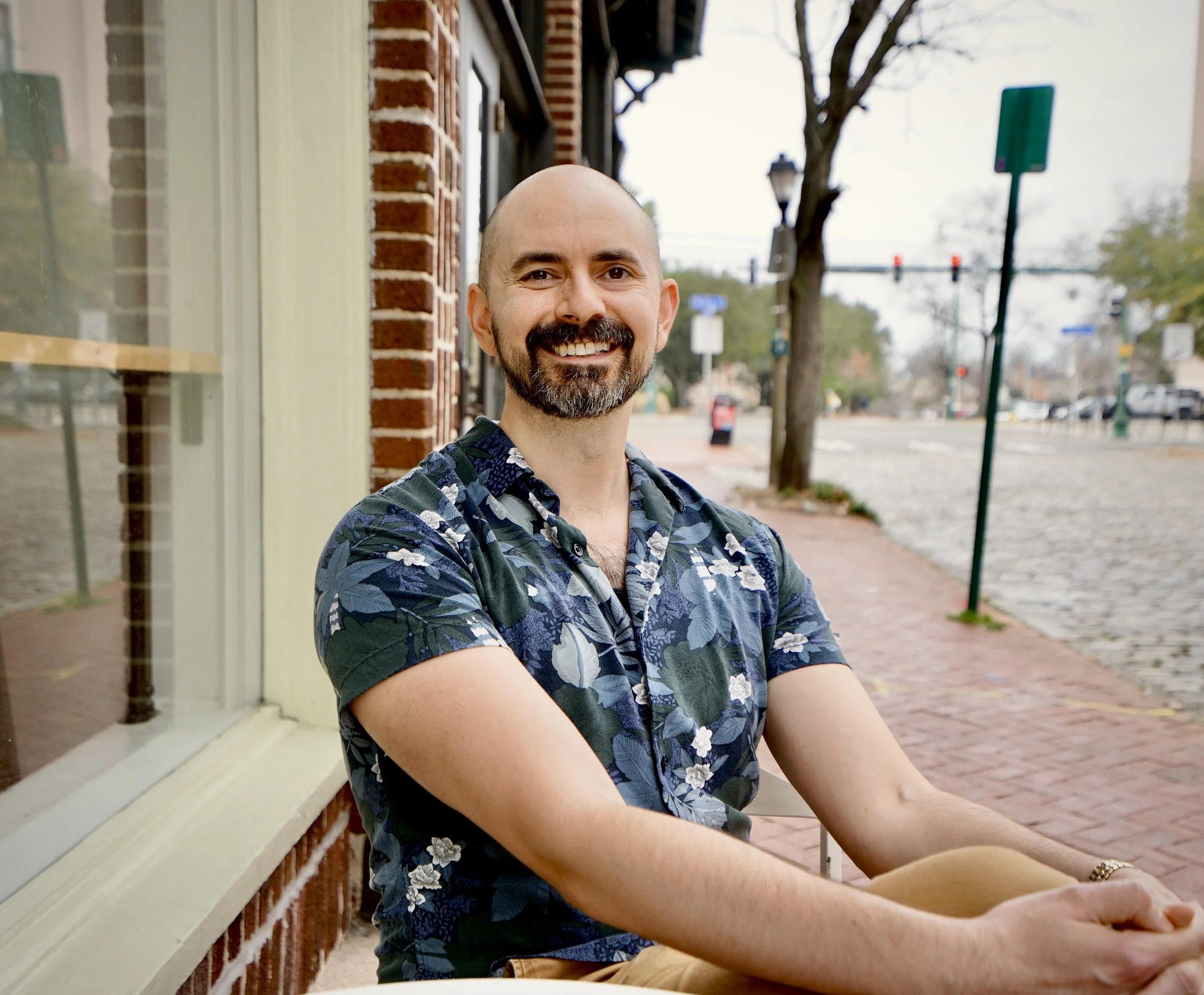 A smiling man with a beard and shaved head wearing a blue floral patterned shirt, sitting outdoors on a sidewalk next to a brick building, with trees, traffic lights, and street signs visible in the background.