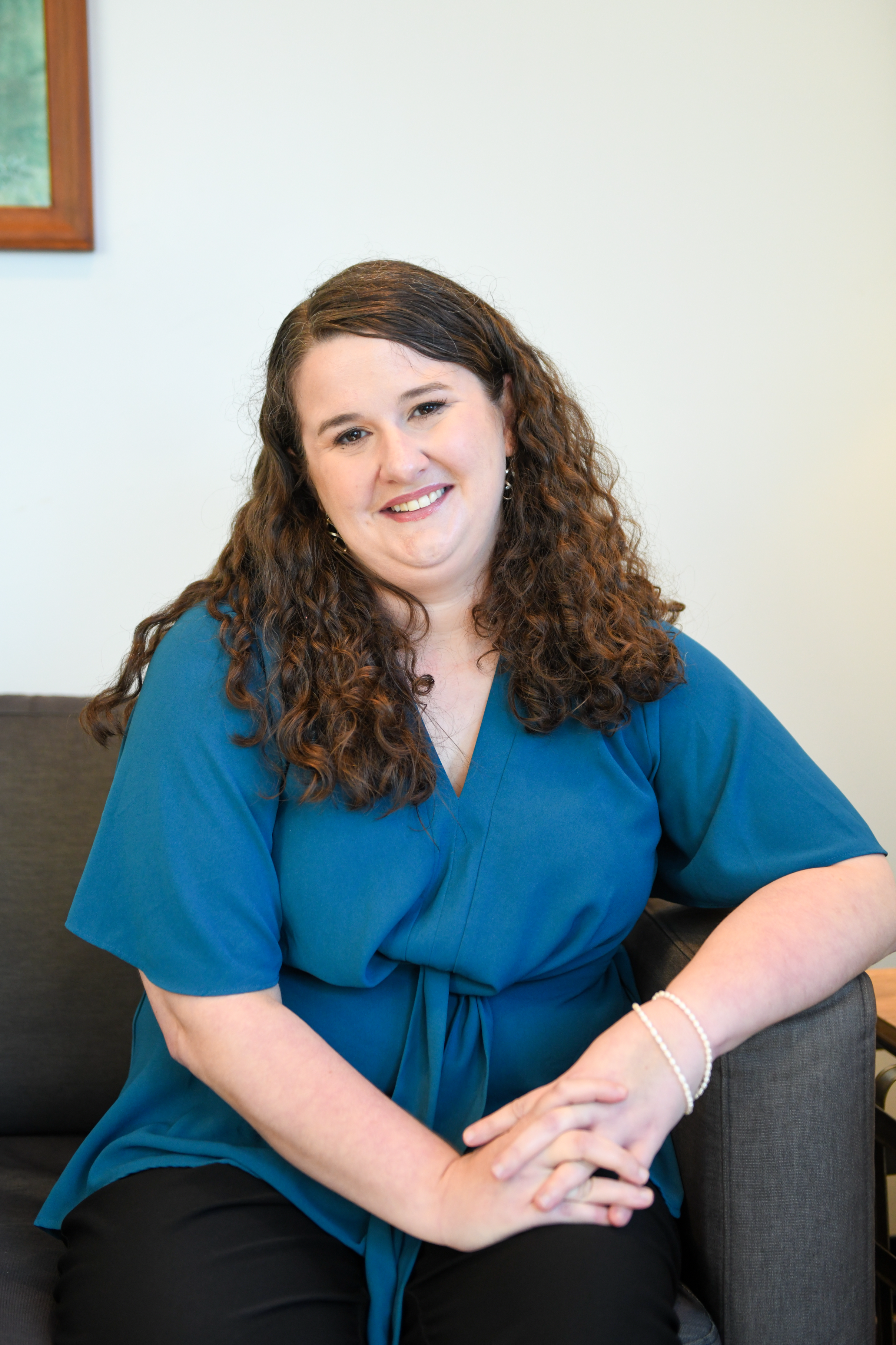 A woman with long curly brown hair wearing a teal blouse and black pants, sitting and smiling in front of a white wall with a wooden-framed artwork.