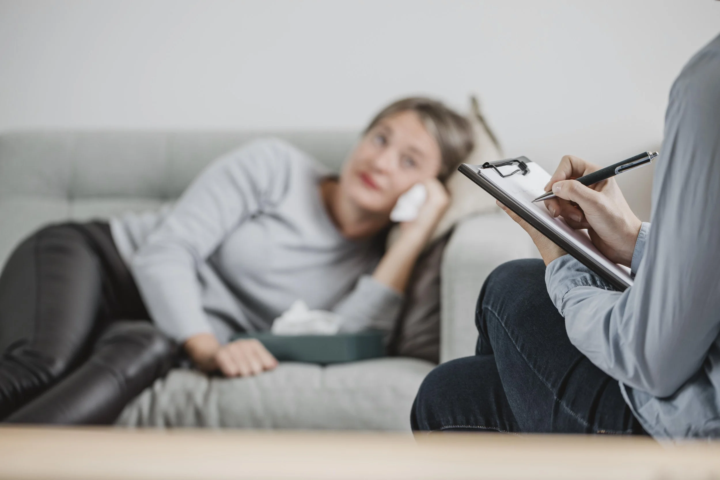 Image of woman laying on a sofa holding a tissue. A person with a clipboard is in the foreground, taking notes.