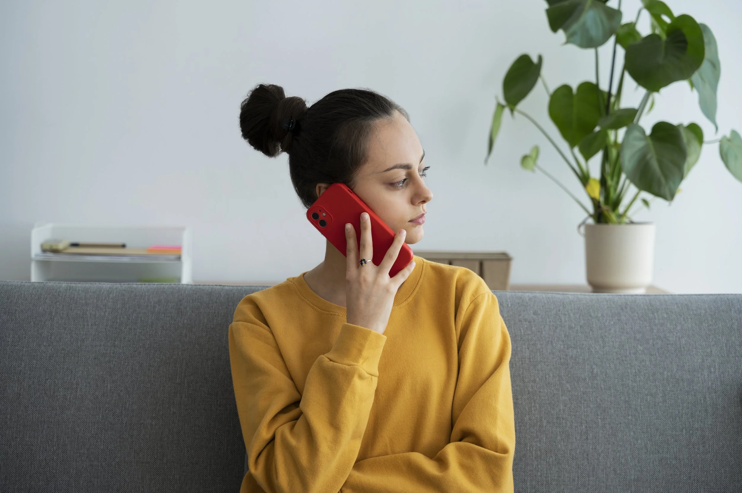 Image of a young woman in a yellow sweatshirt holding a red cellphone to her ear, presumably making a call.