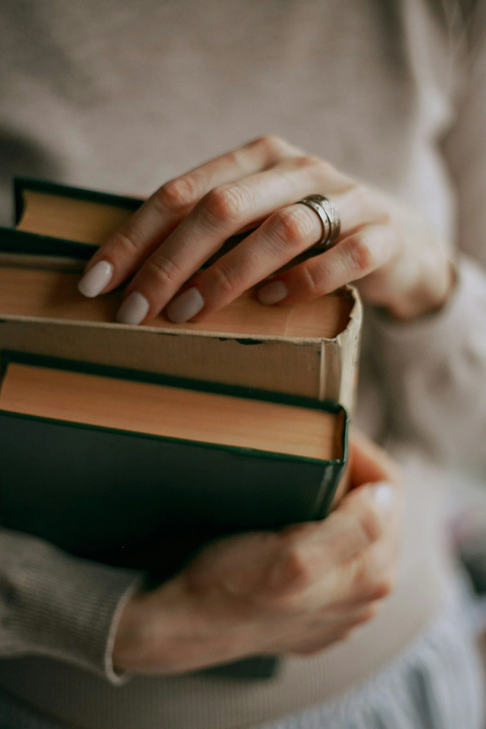 A person holding a stack of three books with one hand, wearing a silver ring on their finger, with a blurred background.