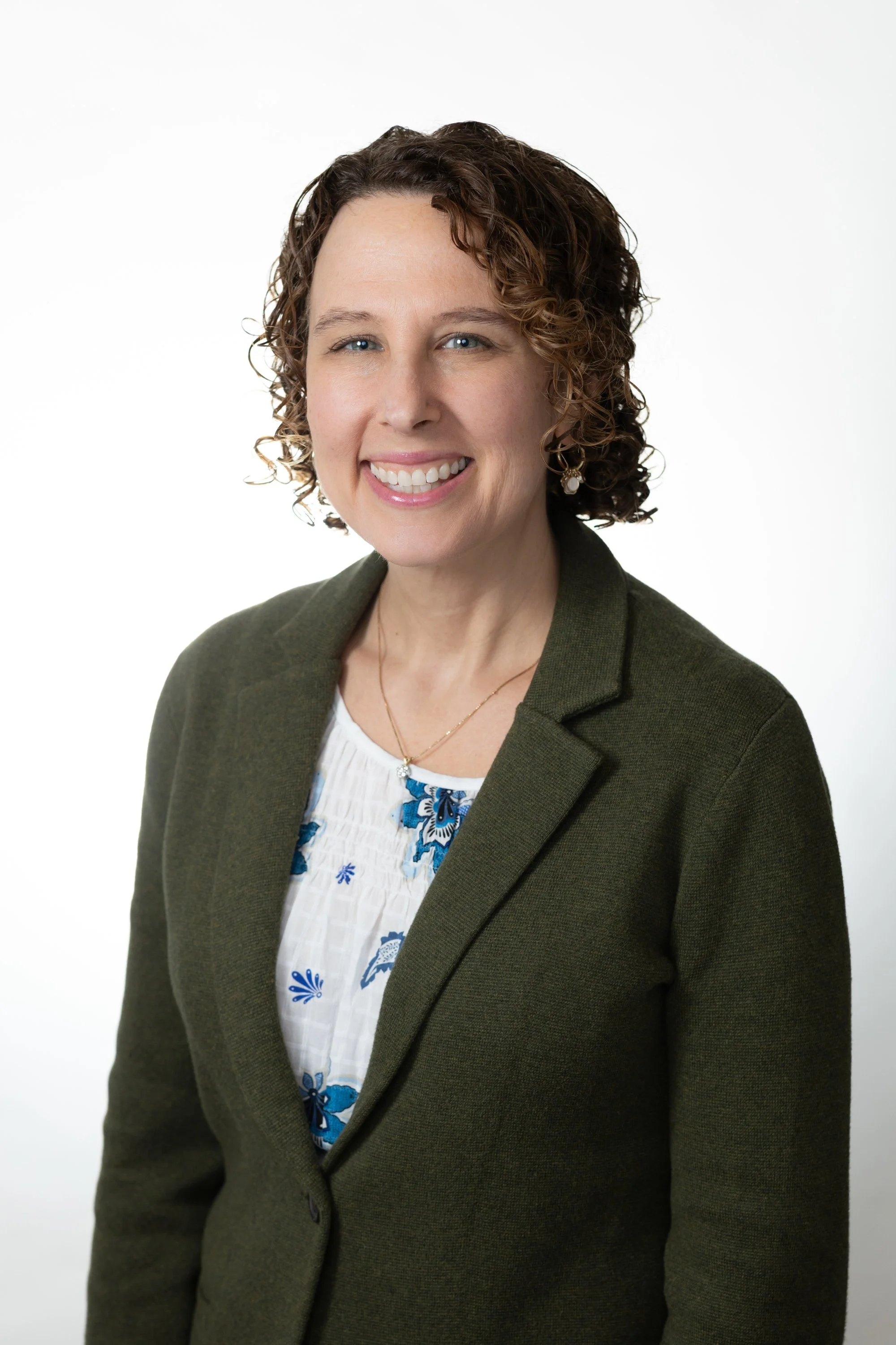 A smiling woman with curly brown hair, wearing a dark green blazer over a white blouse with blue floral embroidery, and earrings, against a plain white background.