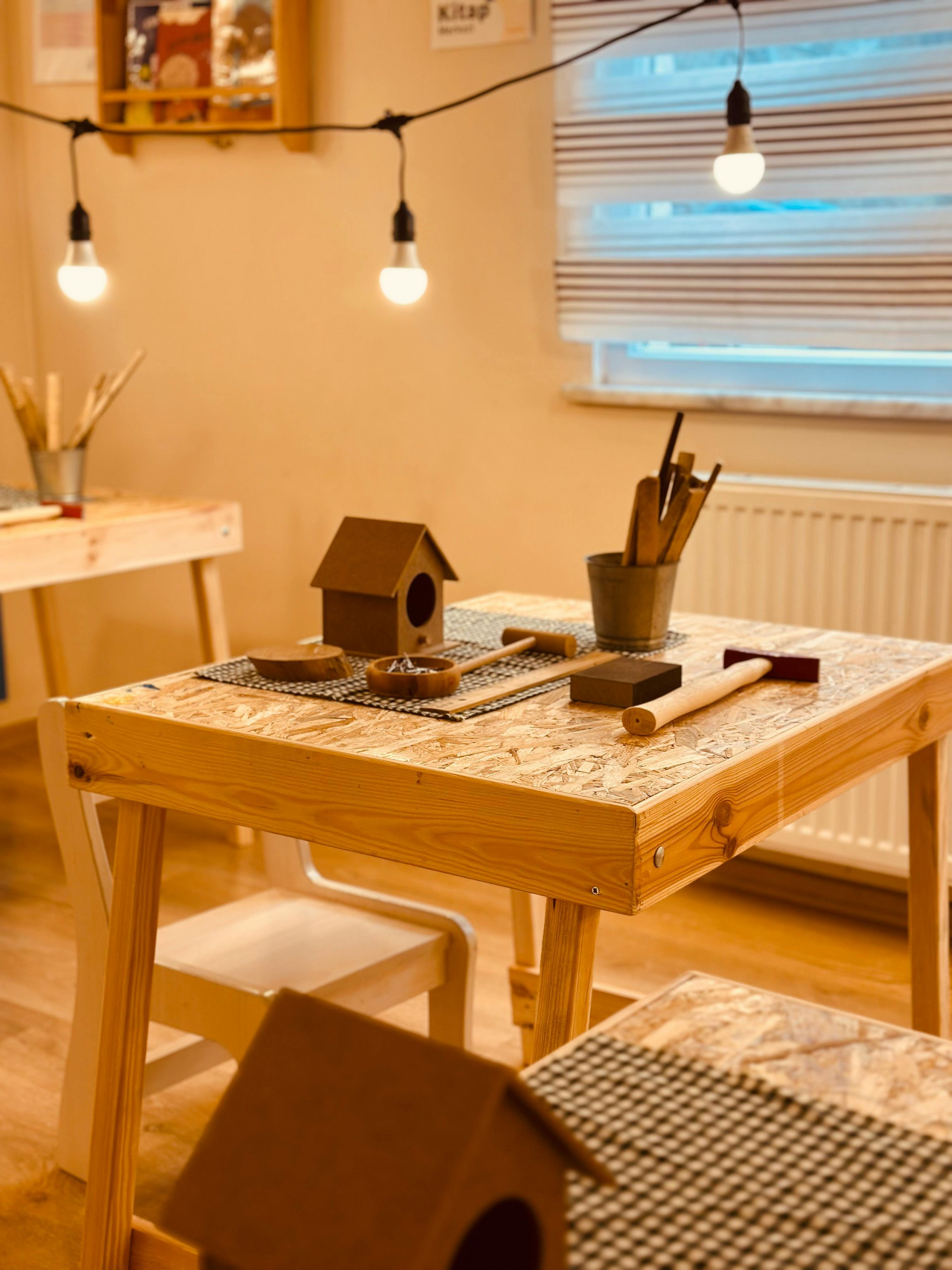 A cozy room with a wooden table set up for arts and crafts, featuring a birdhouse, wooden sticks, and other craft supplies. String lights hang overhead, and a window with closed blinds is visible in the background.